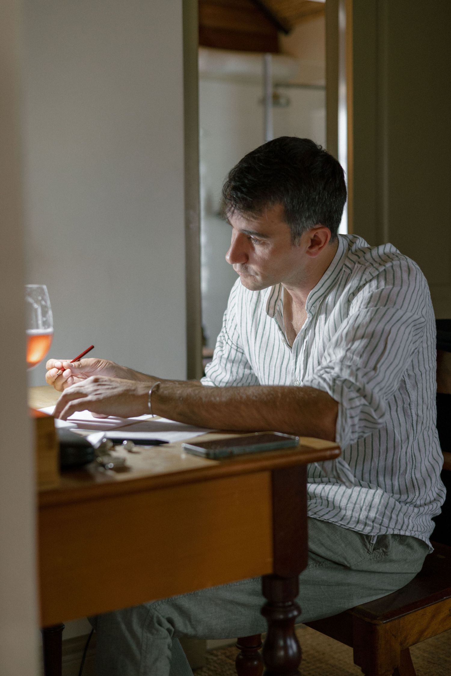 groom in striped shirt writing at wooden desk with pencil, glass of wine nearby, in a dimly lit room.
