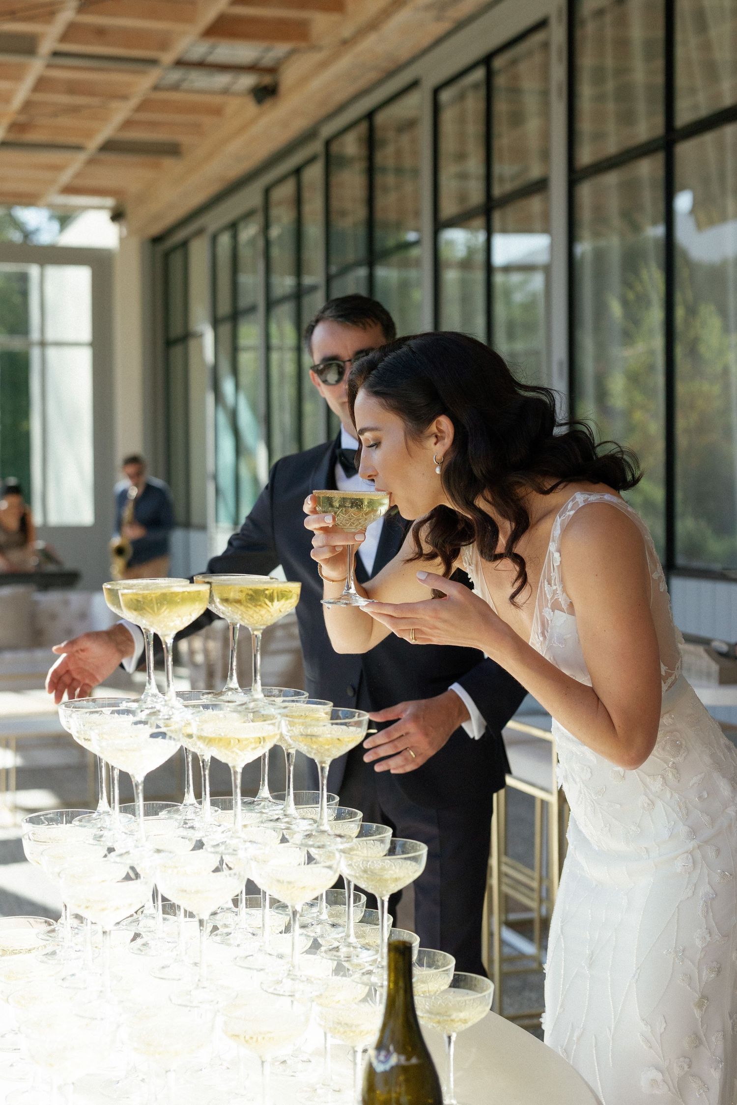 Bride sipping champagne from coupe glass at wedding reception champagne tower display.