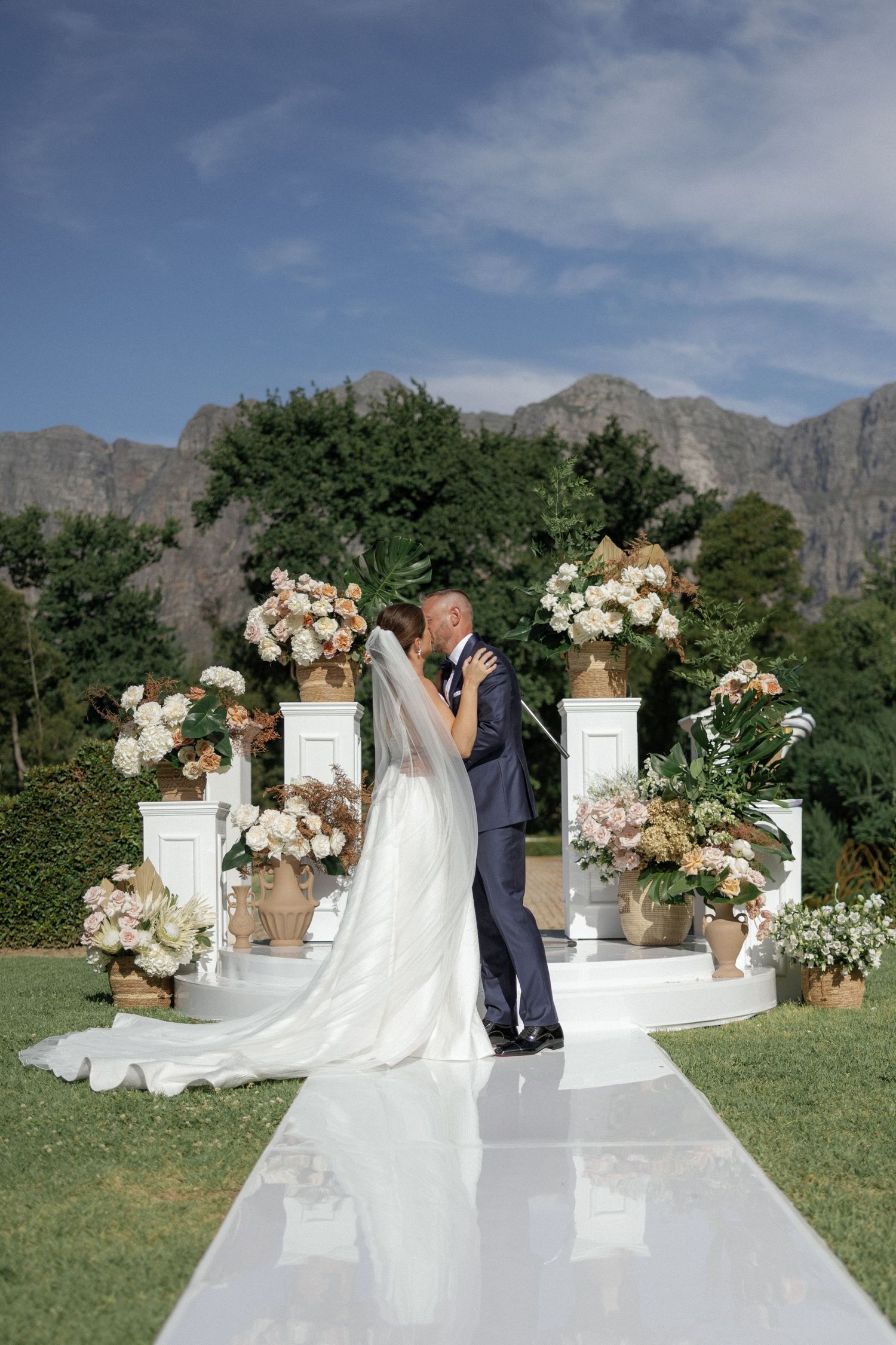Bride and groom share first kiss at outdoor wedding ceremony with floral arrangements and mountain backdrop.