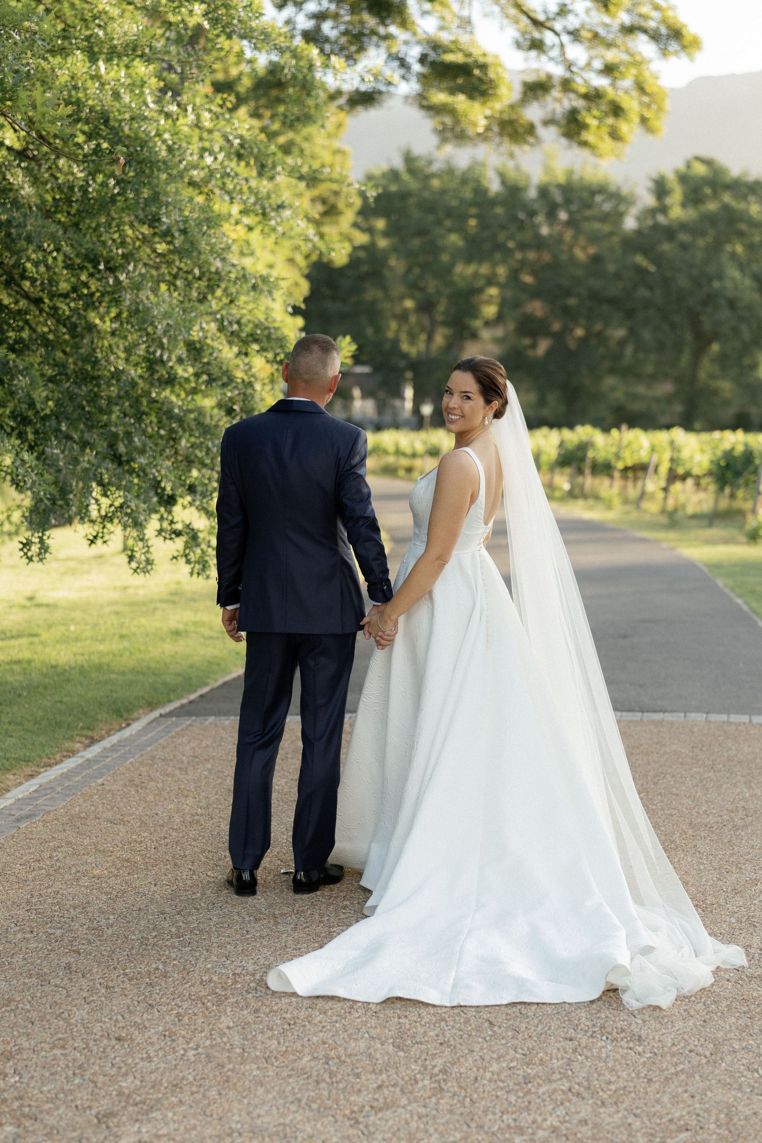 Bride in white gown and veil holds hands with groom in navy suit, walking path through vineyard.
