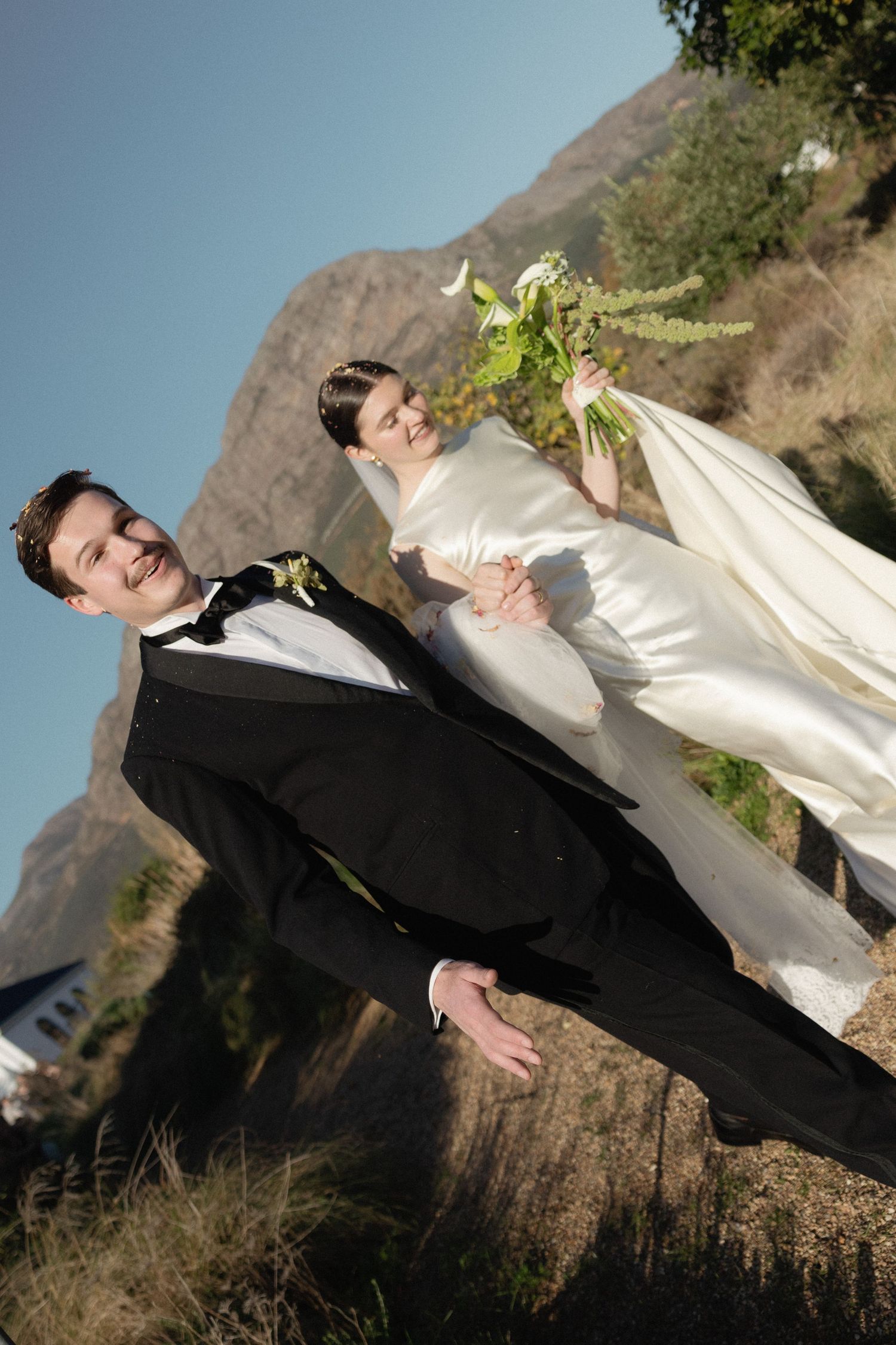 Bride and groom pose outdoors on their wedding day with mountain backdrop, groom in black tuxedo, bride in white gown holding bouquet.