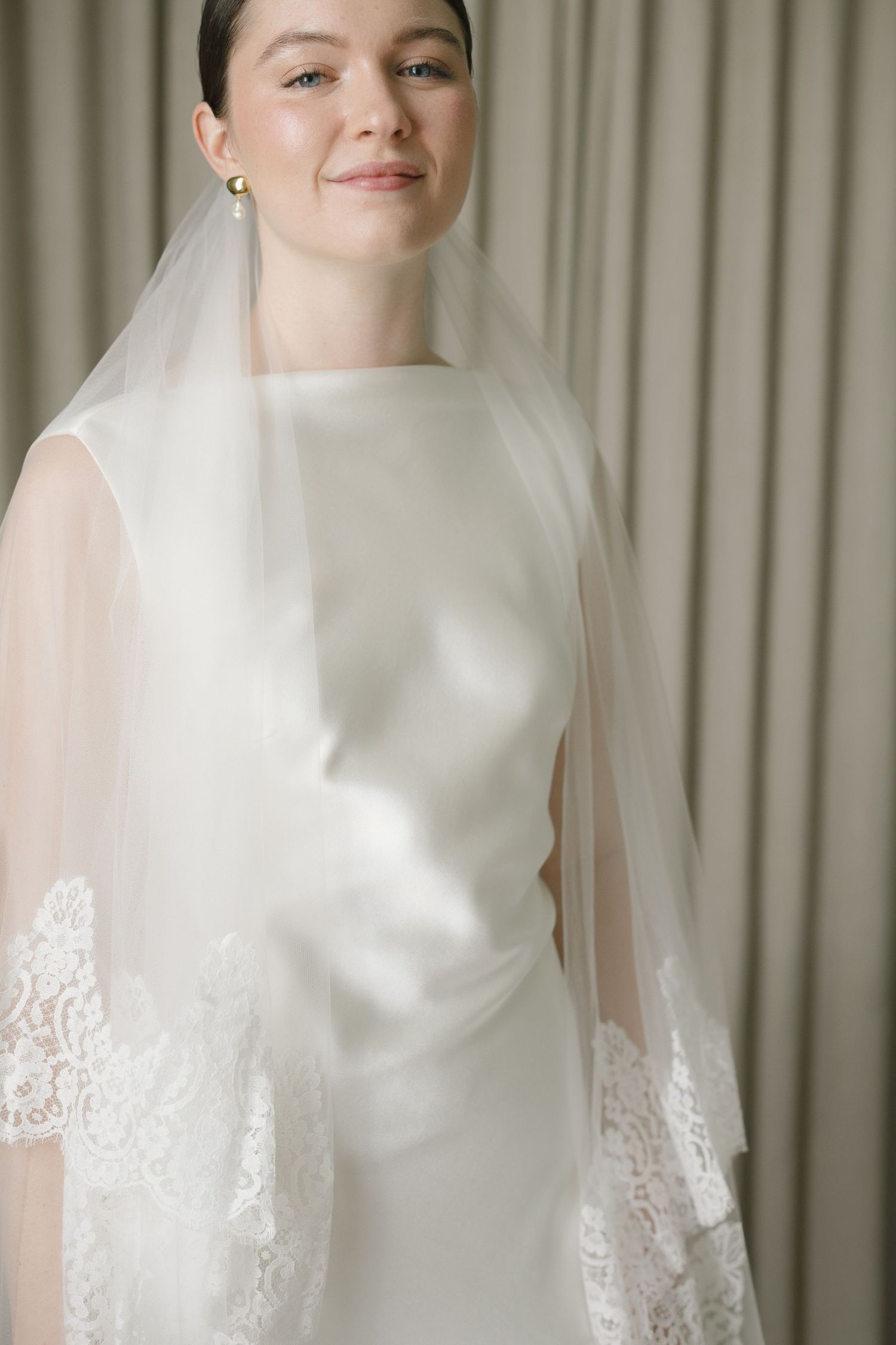 Bride wearing a sleek white satin wedding gown with a lace-trimmed veil and gold drop earrings, posing elegantly.