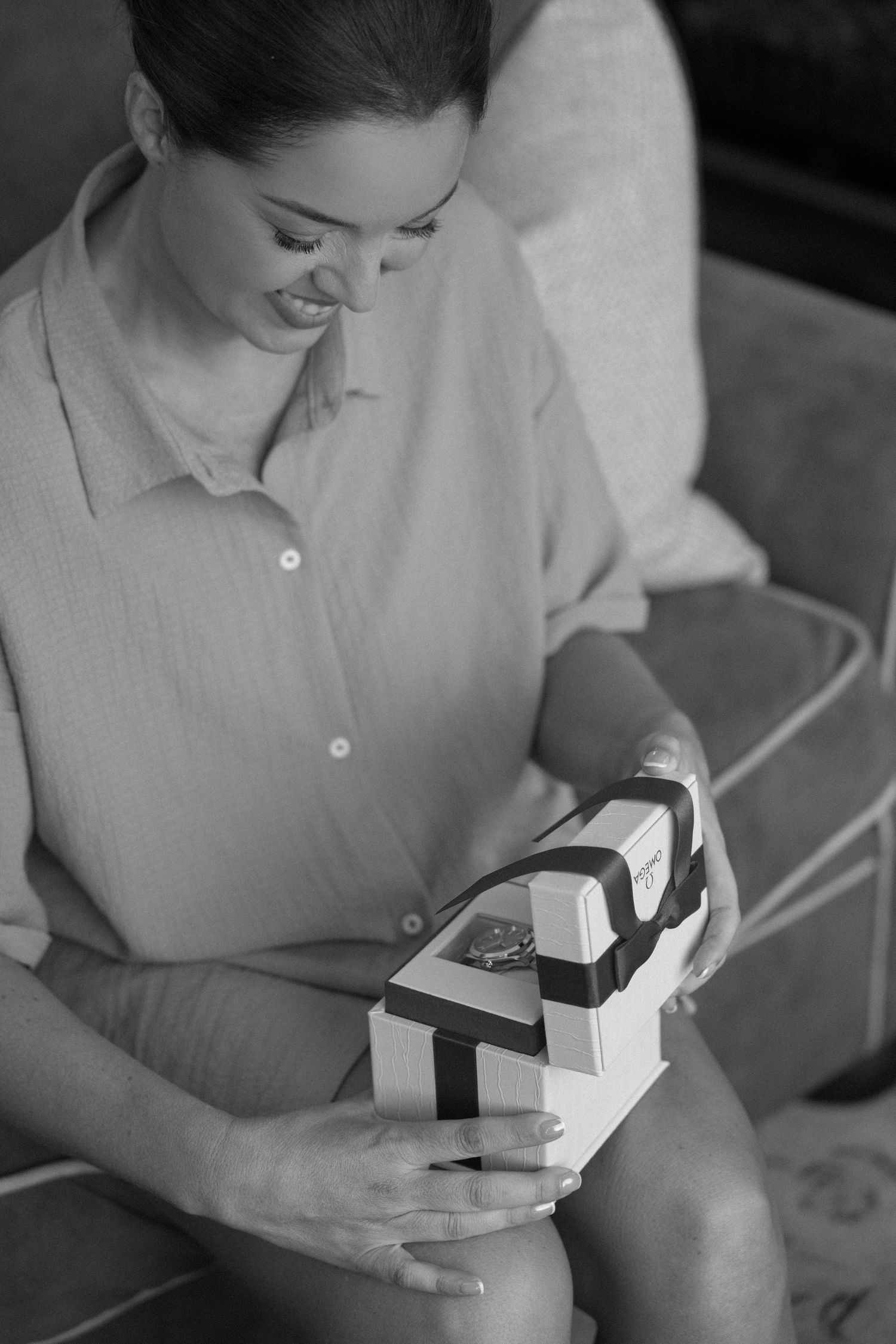 Woman smiling while opening a gift box with a ribbon, sitting on a couch in a casual shirt, black and white photo.