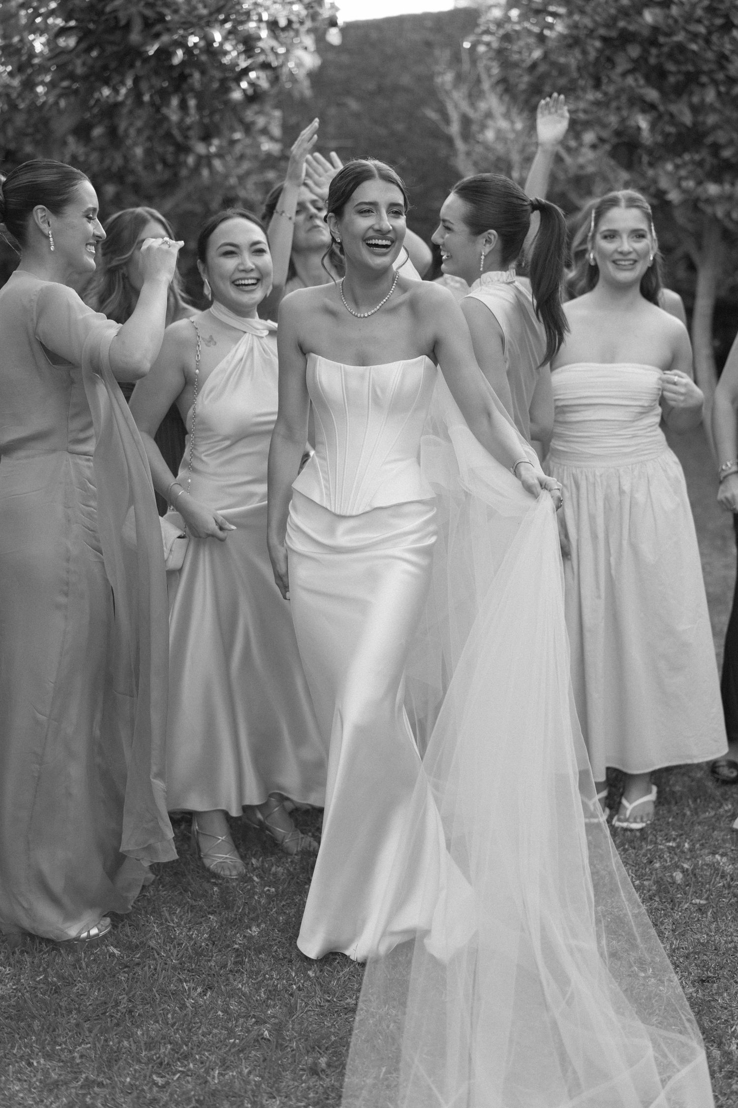 Black and white photo of a smiling bride in a strapless gown with bridesmaids in matching dresses celebrating outdoors.