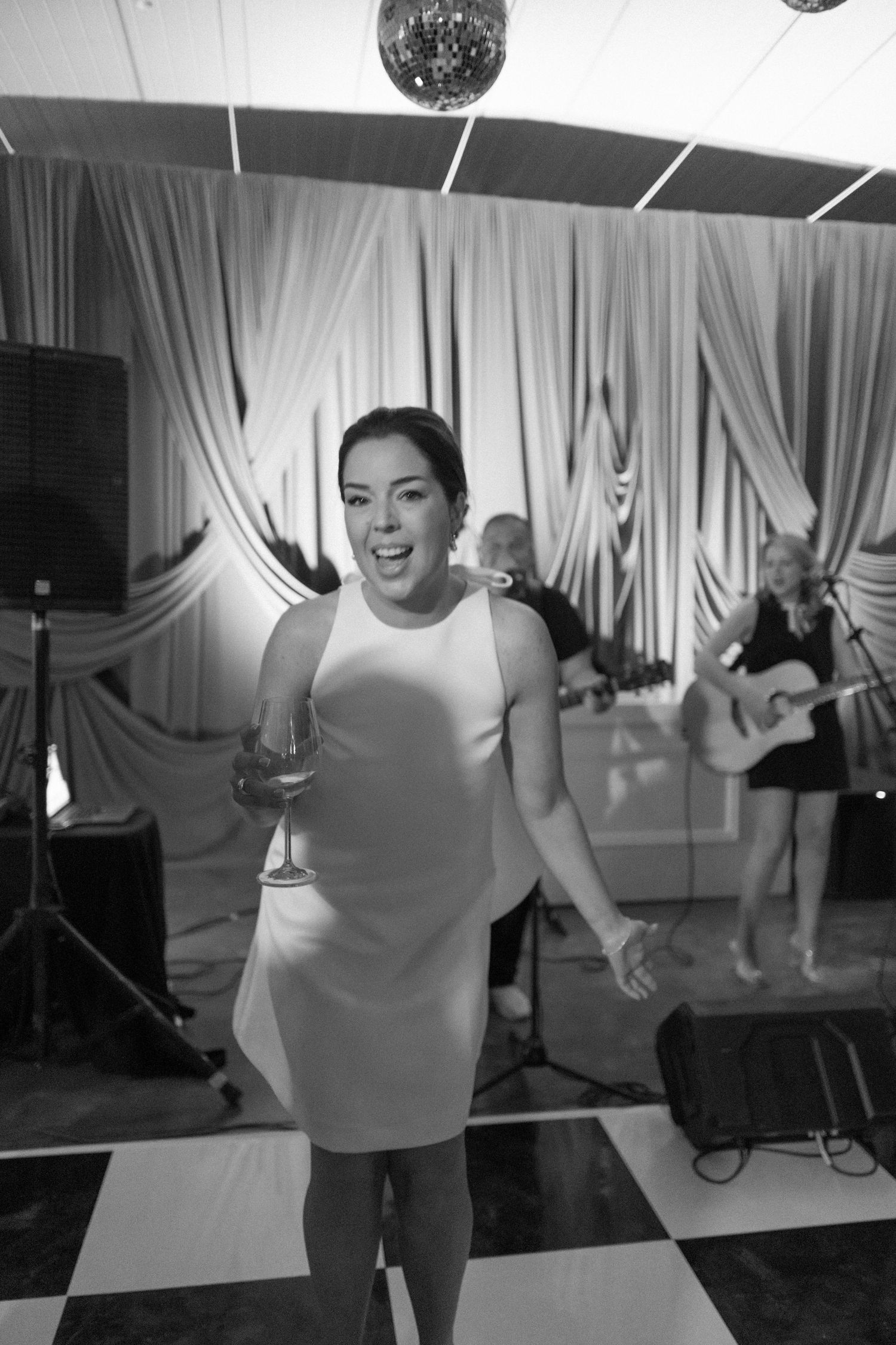 Woman in white dress dancing with wine glass at wedding reception with live band on stage.