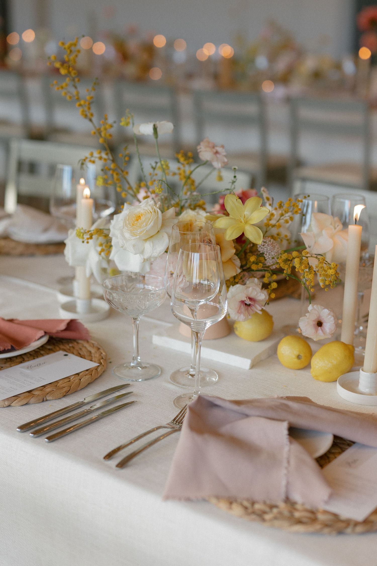 Elegant wedding table setting with white roses, yellow mimosa, lemons, candles, crystal glasses, and blush pink linen napkins.