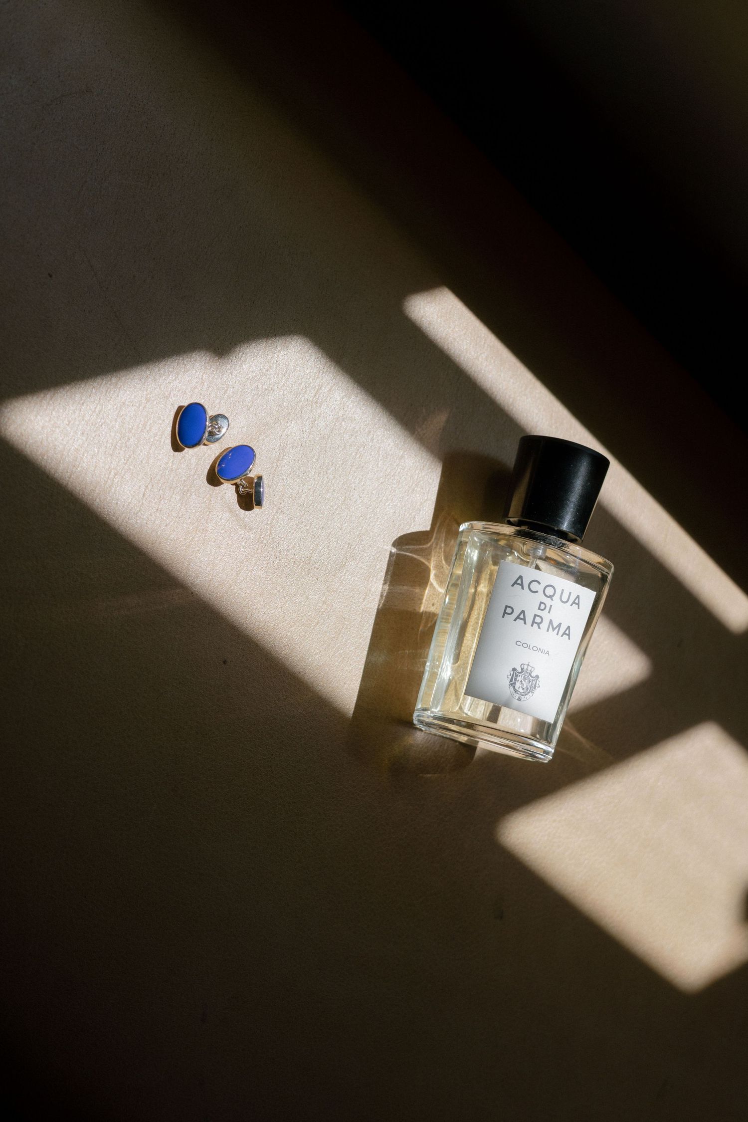 Acqua di Parma perfume bottle with blue heart earrings in dramatic sunlight shadows.