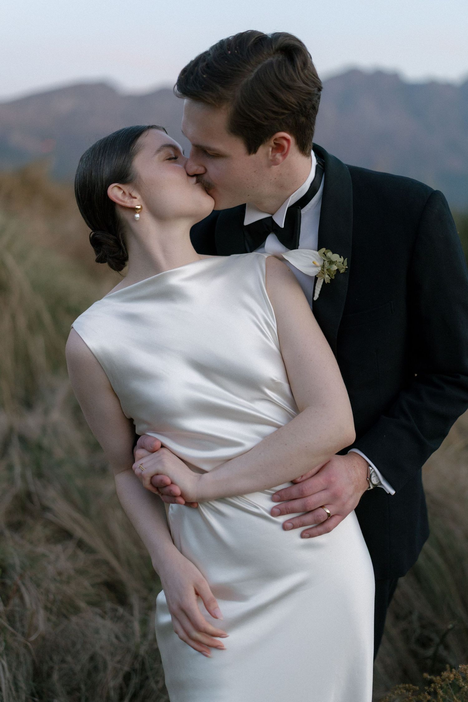 Newlyweds share a passionate kiss outdoors, bride in elegant white satin dress, groom in black tuxedo.