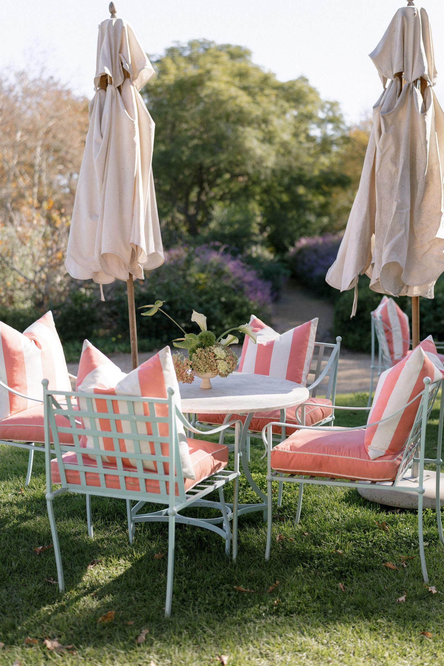 Elegant outdoor garden seating with white metal chairs, coral striped cushions, and closed cream umbrellas.