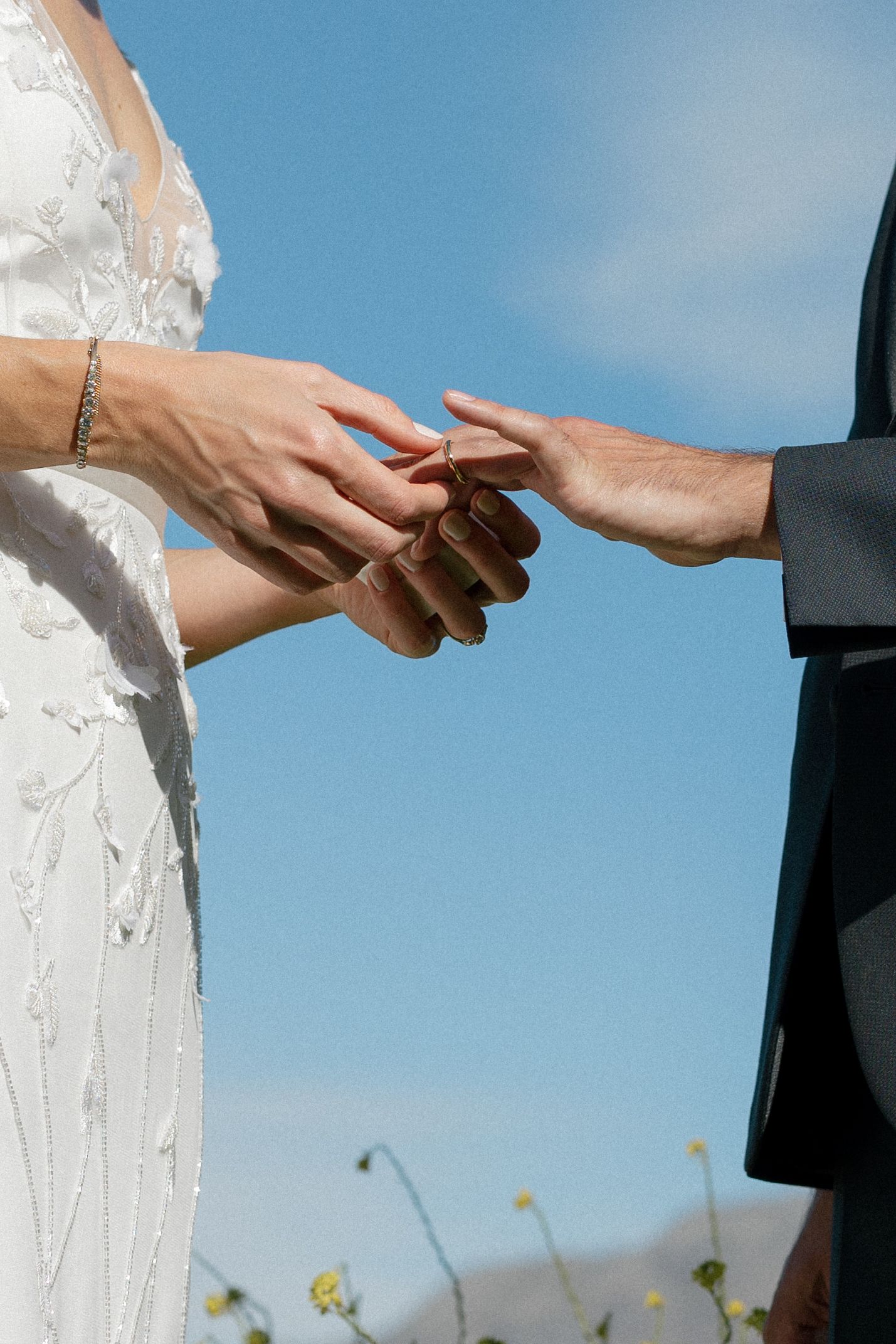 Close-up of groom placing wedding ring on brides hand against a clear blue sky at outdoor ceremony.