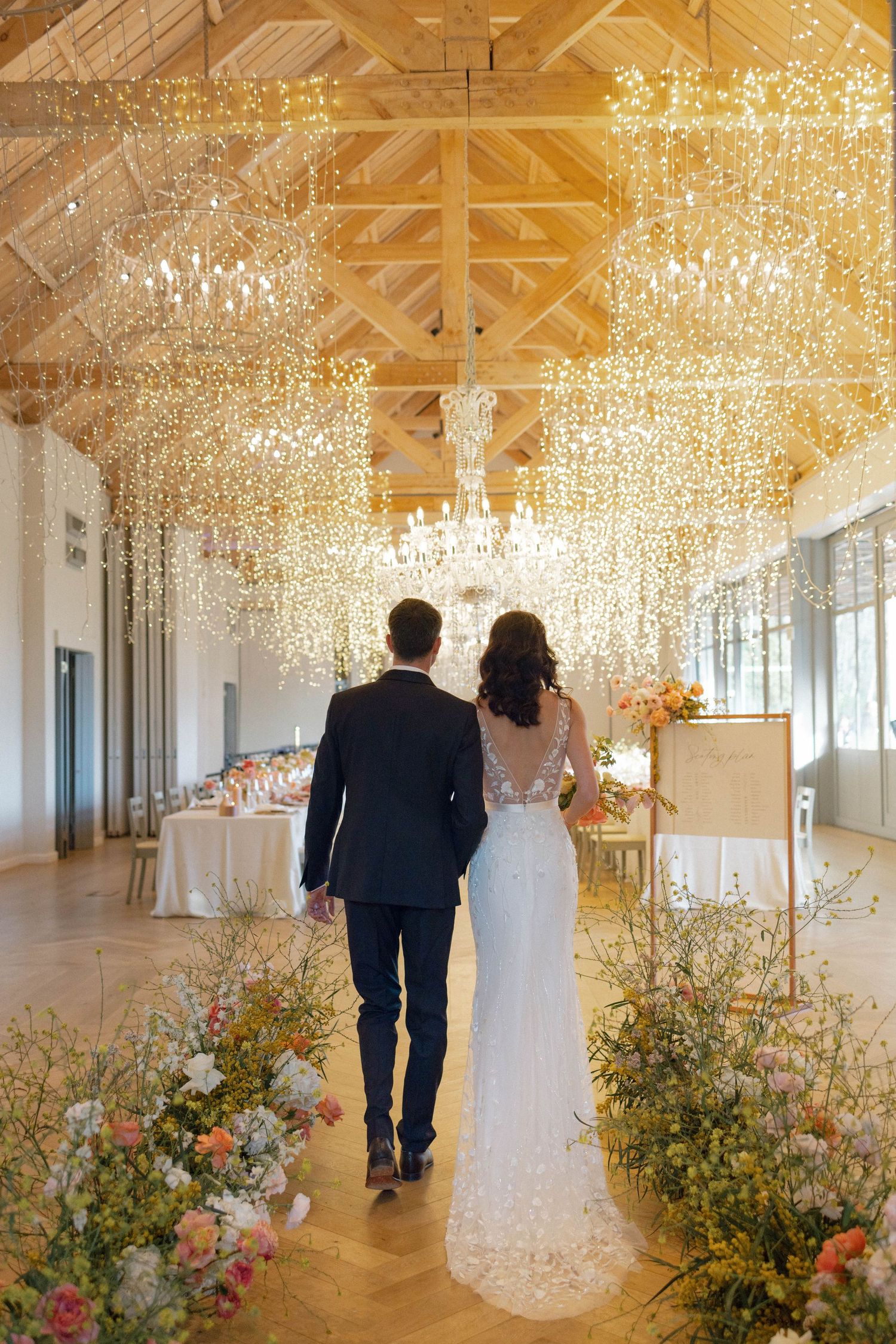 Bride and groom walking down aisle under sparkling chandeliers in elegant barn venue.