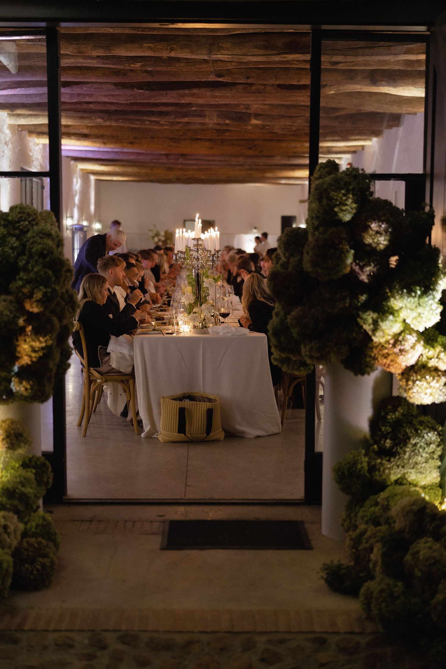 Elegant candlelit wedding party with lush green floral arrangements framing guests seated at a long white-draped table.