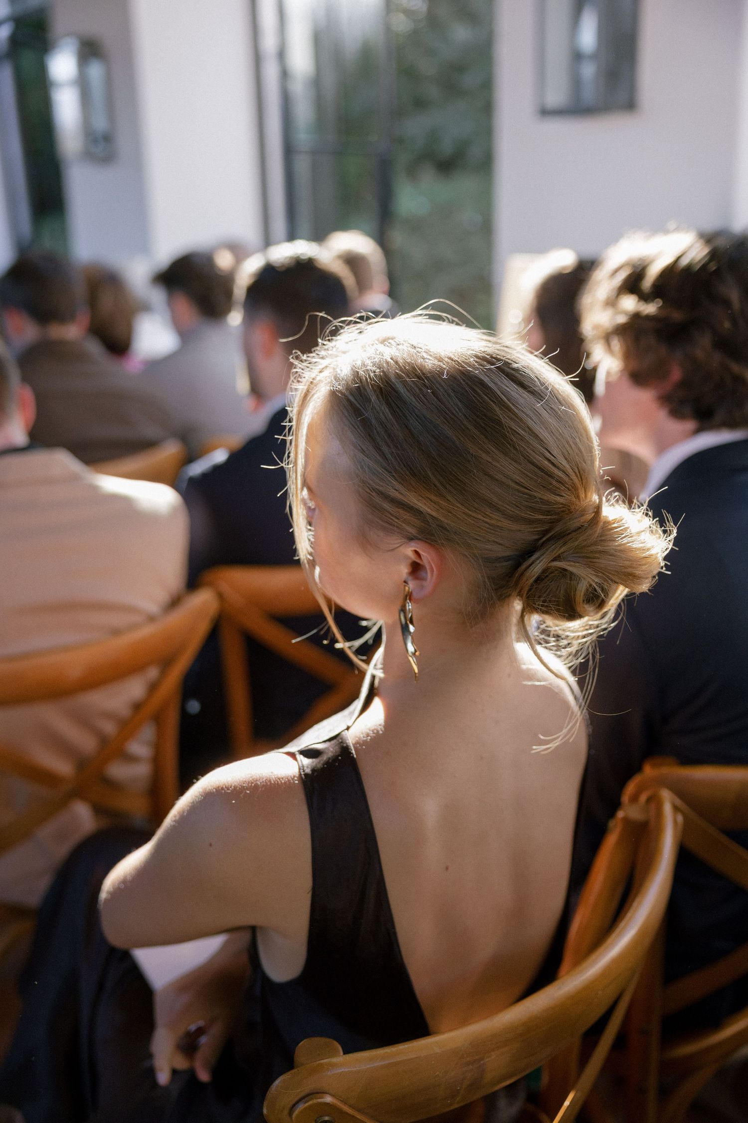 Wedding guest with elegant updo and drop earrings in black dress seated at outdoor ceremony.