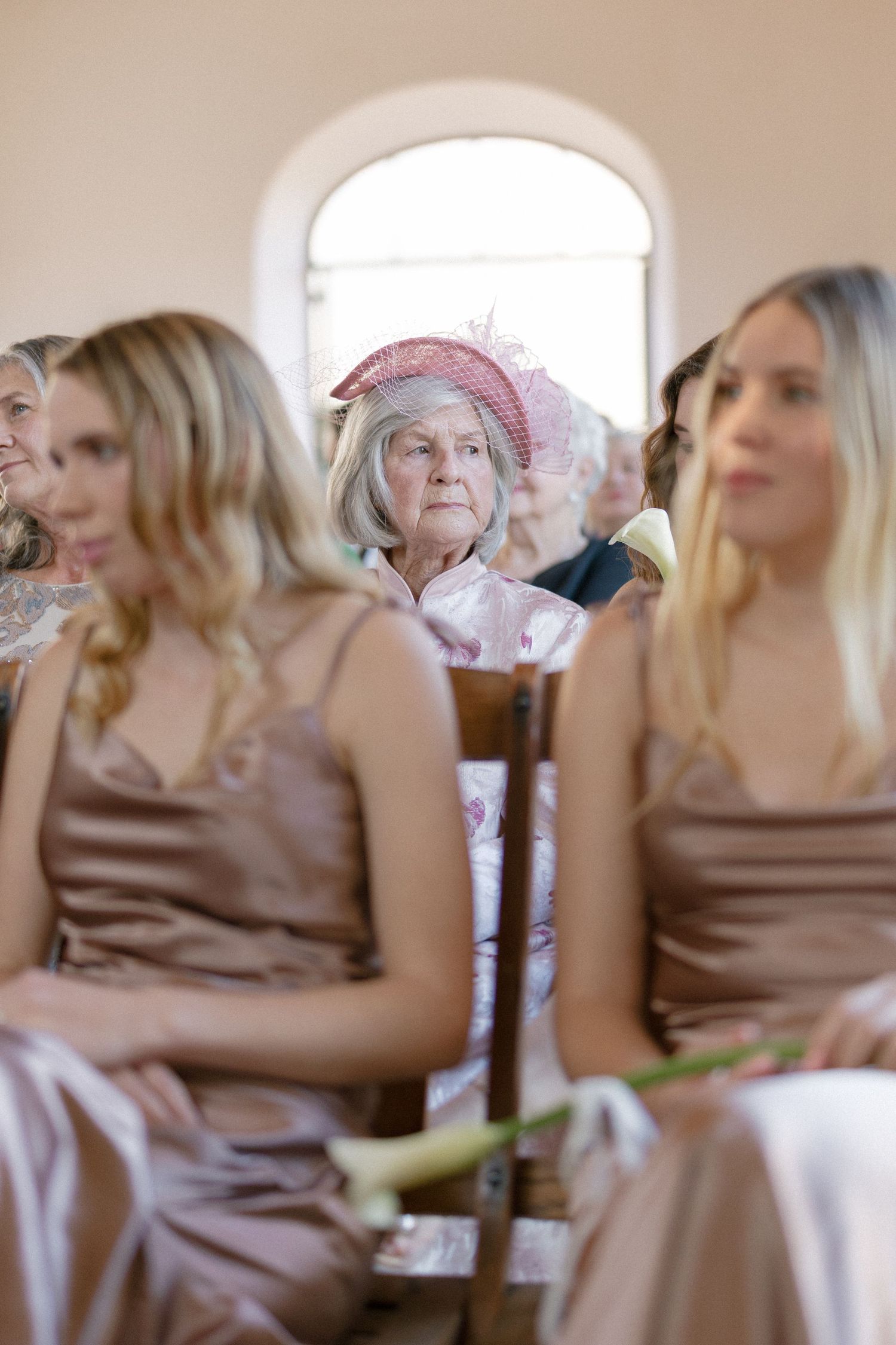 Elderly woman in pink hat watches wedding ceremony seated among bridesmaids in taupe gowns.
