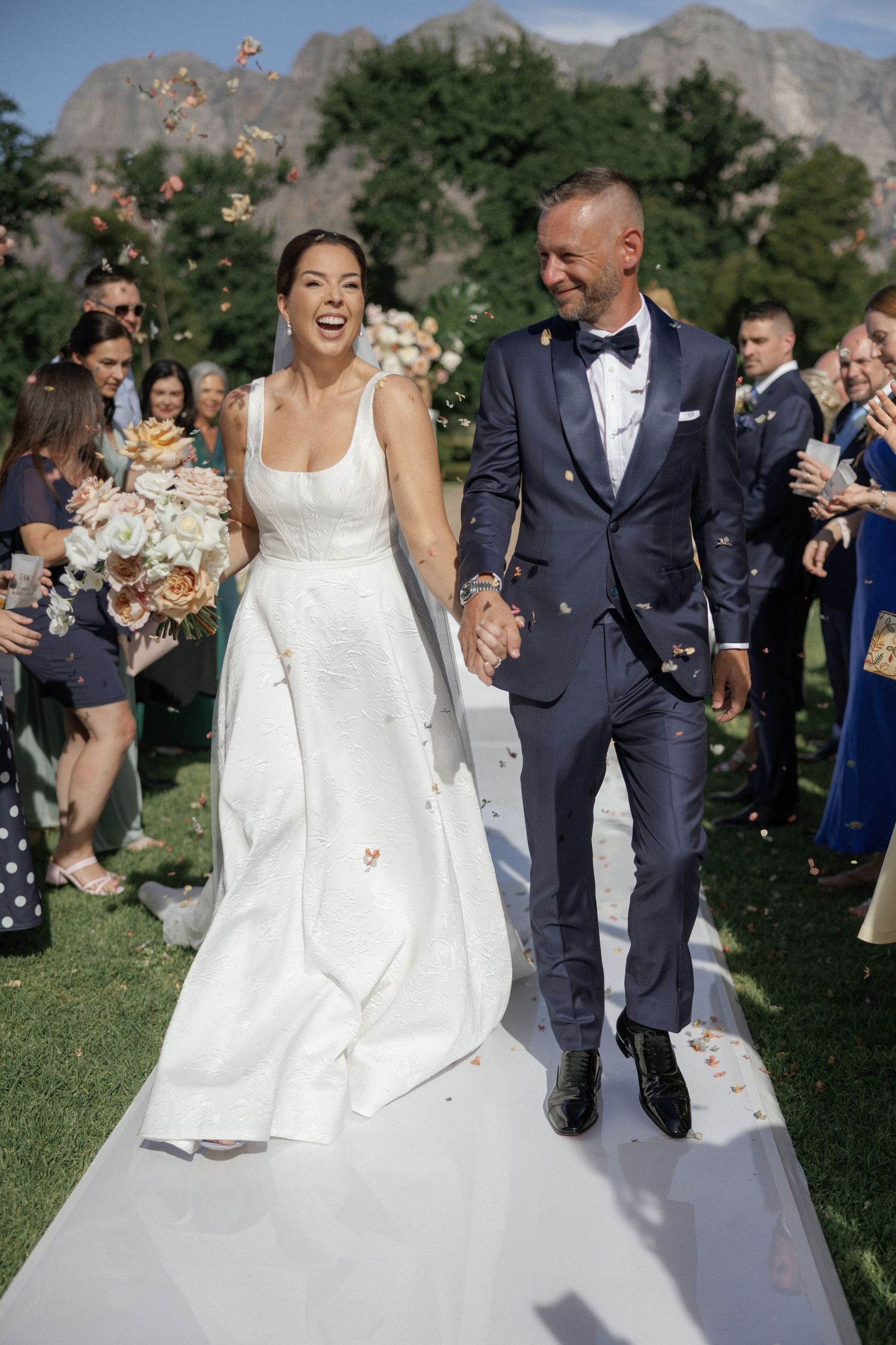 Bride and groom walk down aisle laughing as guests toss confetti at outdoor wedding with mountain backdrop.