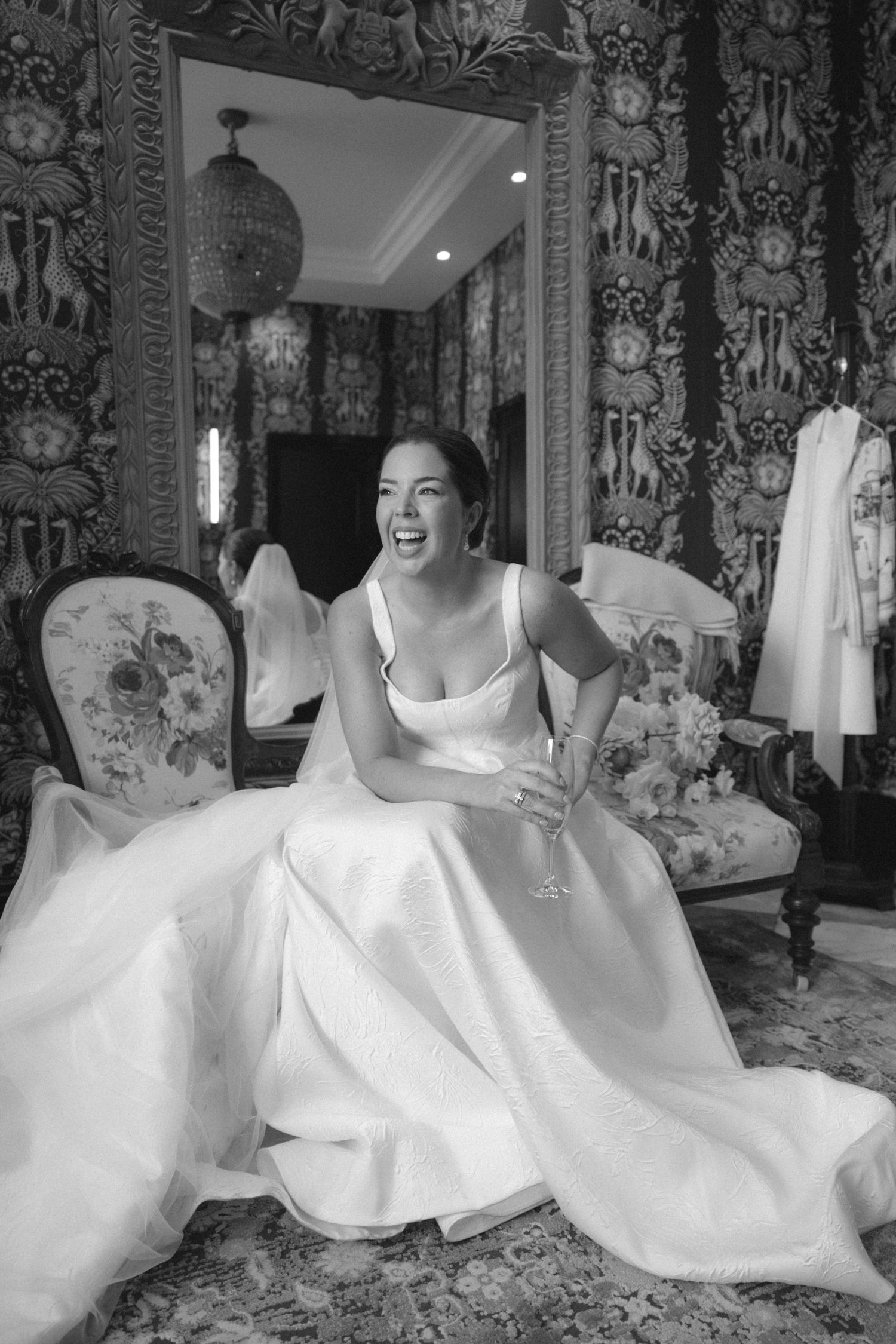 Black and white photo of a laughing bride in a white gown holding a champagne glass in an ornate room.