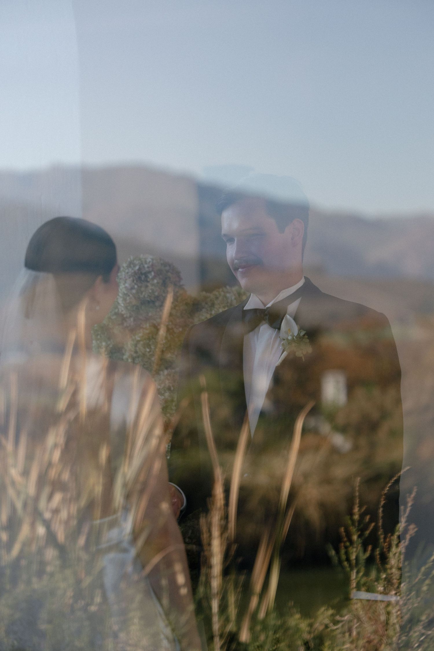 Bride and groom in formal attire reflected through glass with scenic mountain backdrop.