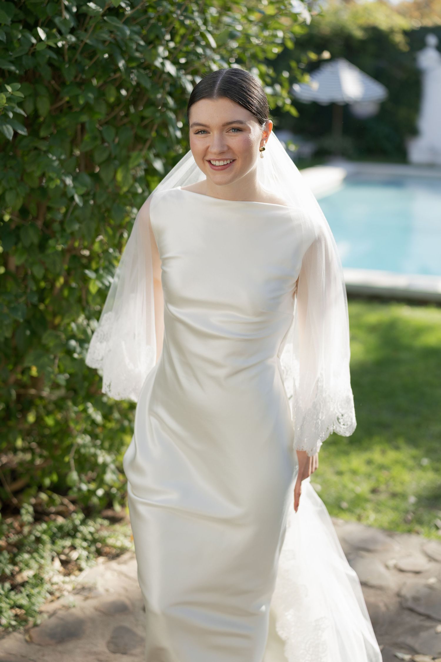 Smiling bride in elegant white satin gown with lace veil poses outdoors near pool and greenery.