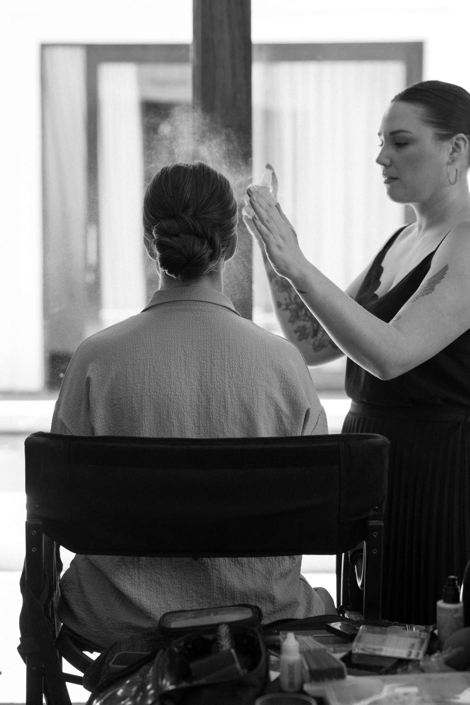Black and white photo of makeup artist applying powder to seated client in director's chair at vanity.