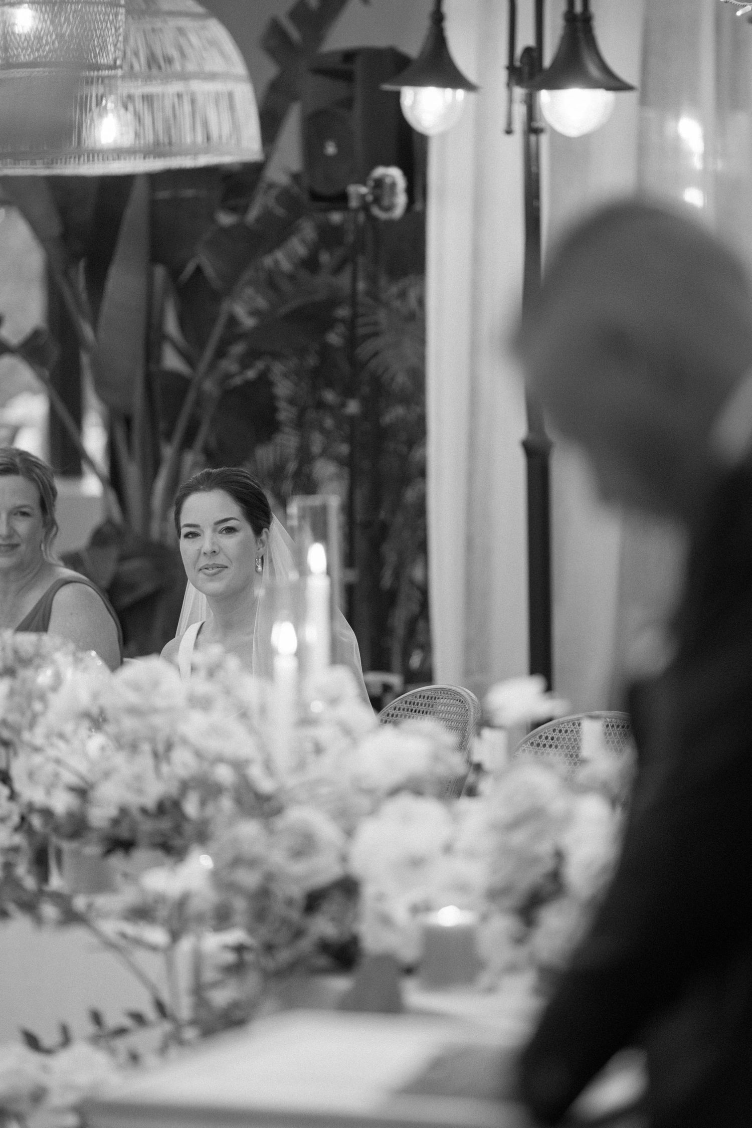 Black and white wedding photo of a bride seated at a floral reception table, gazing thoughtfully amid candlelight.