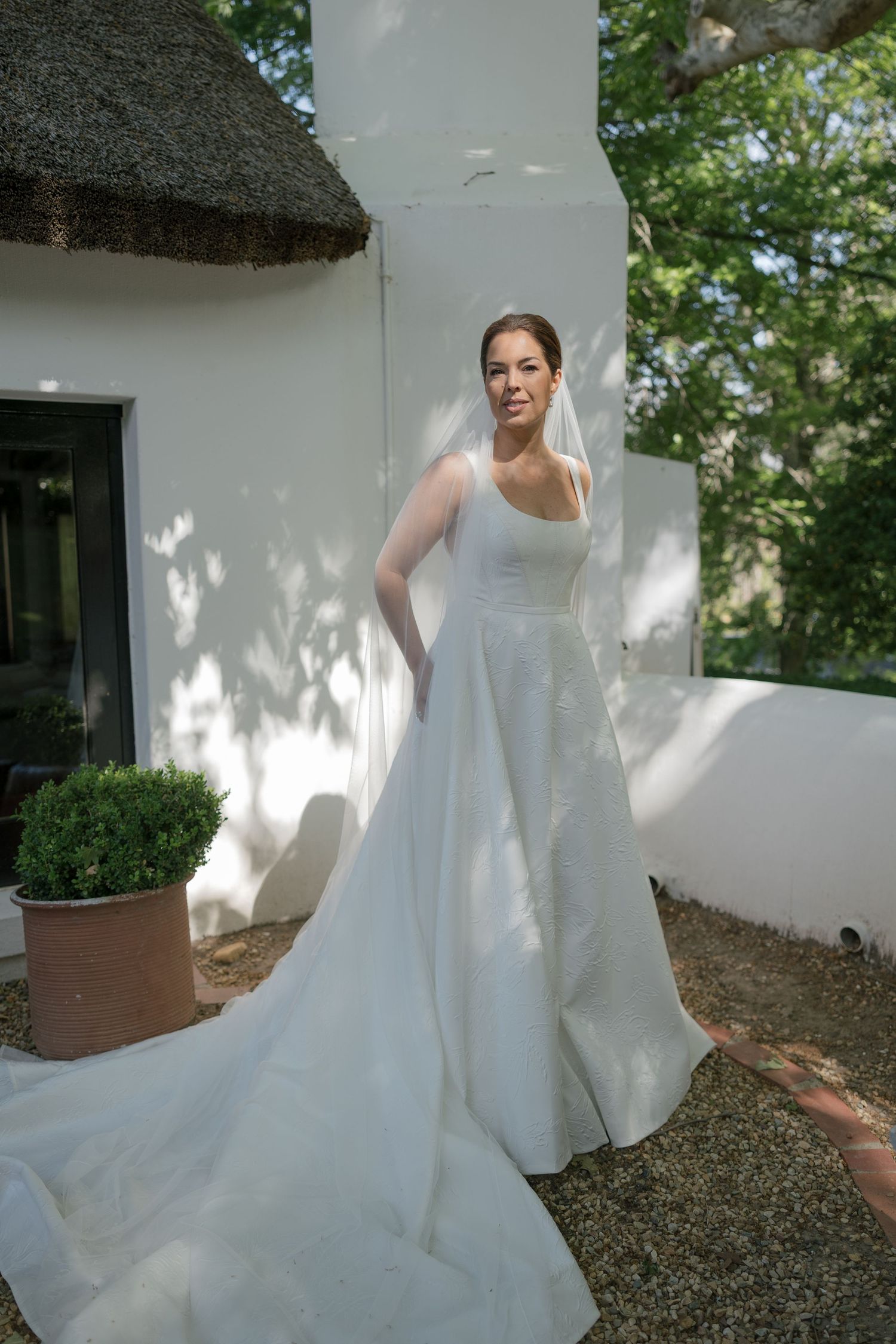 Bride in elegant white A-line wedding gown with long train and veil, posing outdoors by a whitewashed wall.