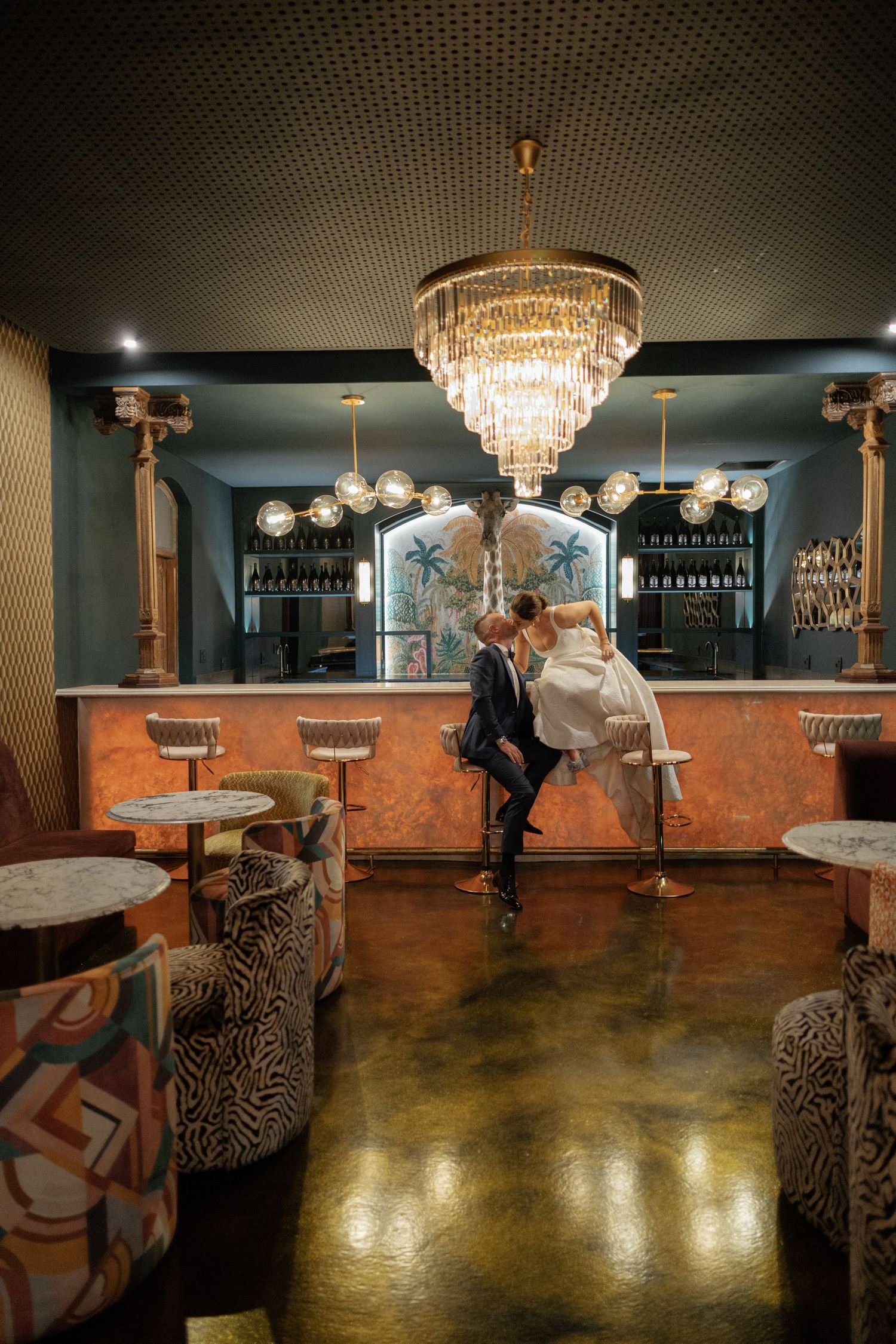 Bride and groom share a romantic moment at an elegant bar with a gold chandelier and Art Deco decor.