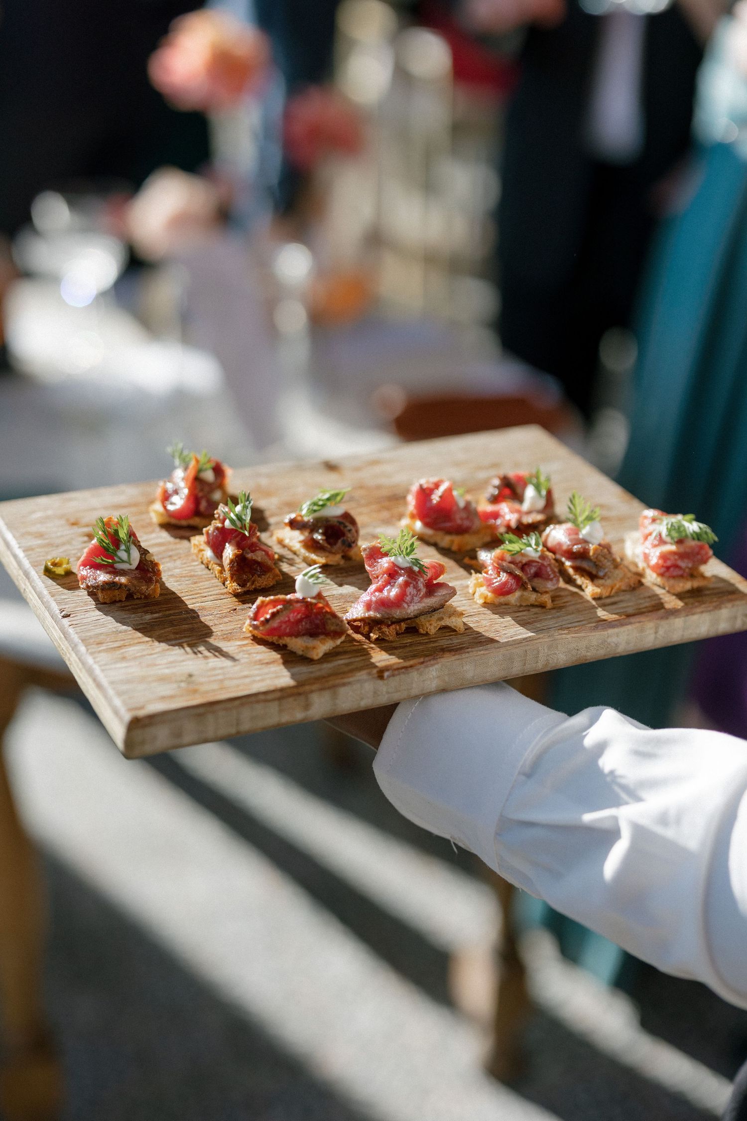Wedding catering server holds wooden board with elegant meat and herb topped crostini appetizers.