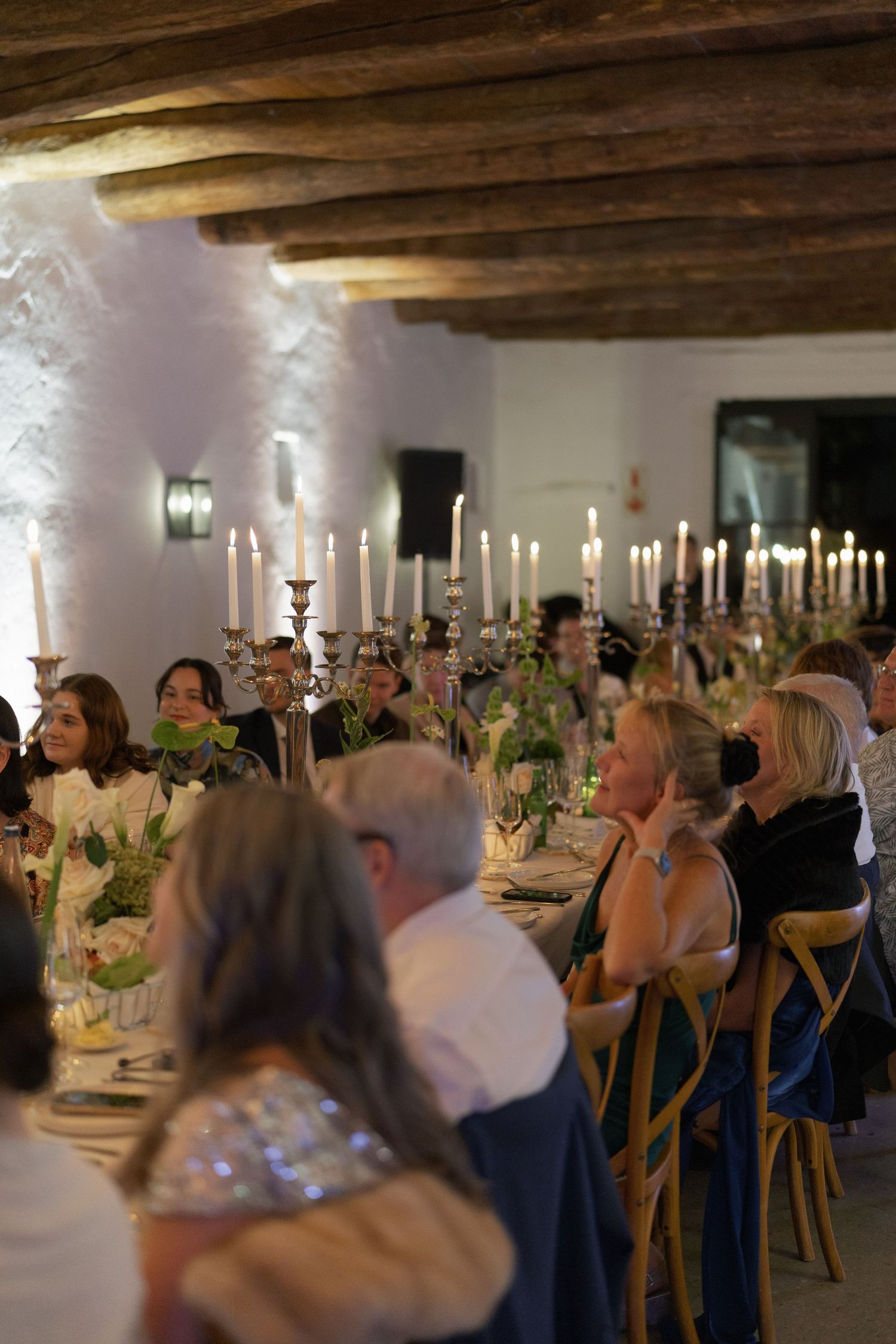 Elegant wedding reception dinner with guests seated at long candlelit tables under rustic wooden beam ceiling.