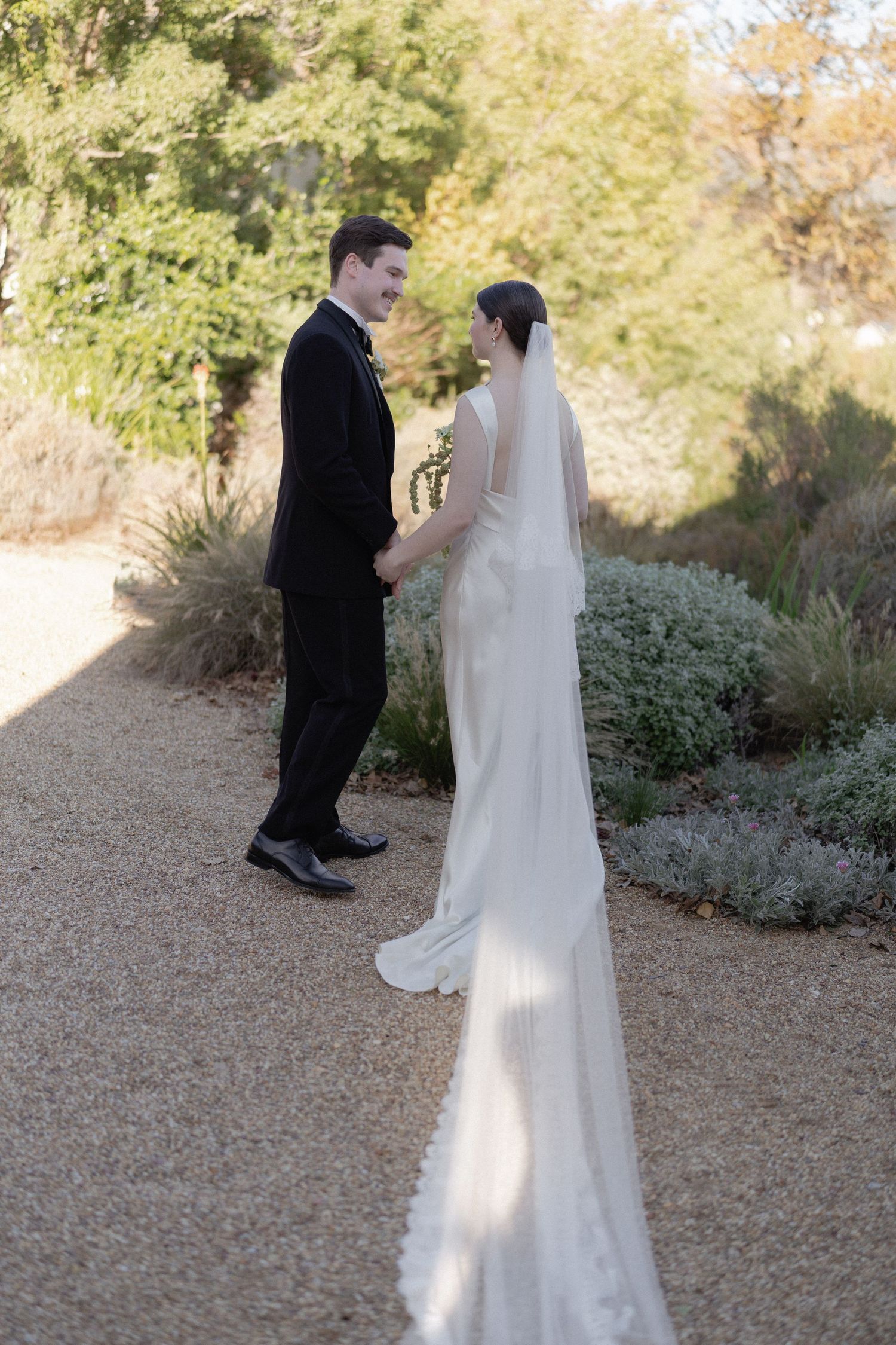 Bride in white gown with lace veil holding hands with groom in black tuxedo on garden gravel path.