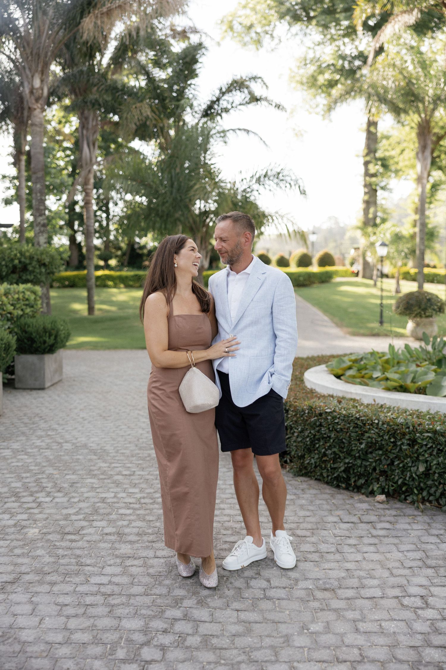 A smiling couple poses on a brick pathway surrounded by lush tropical gardens and palm trees on a sunny day.