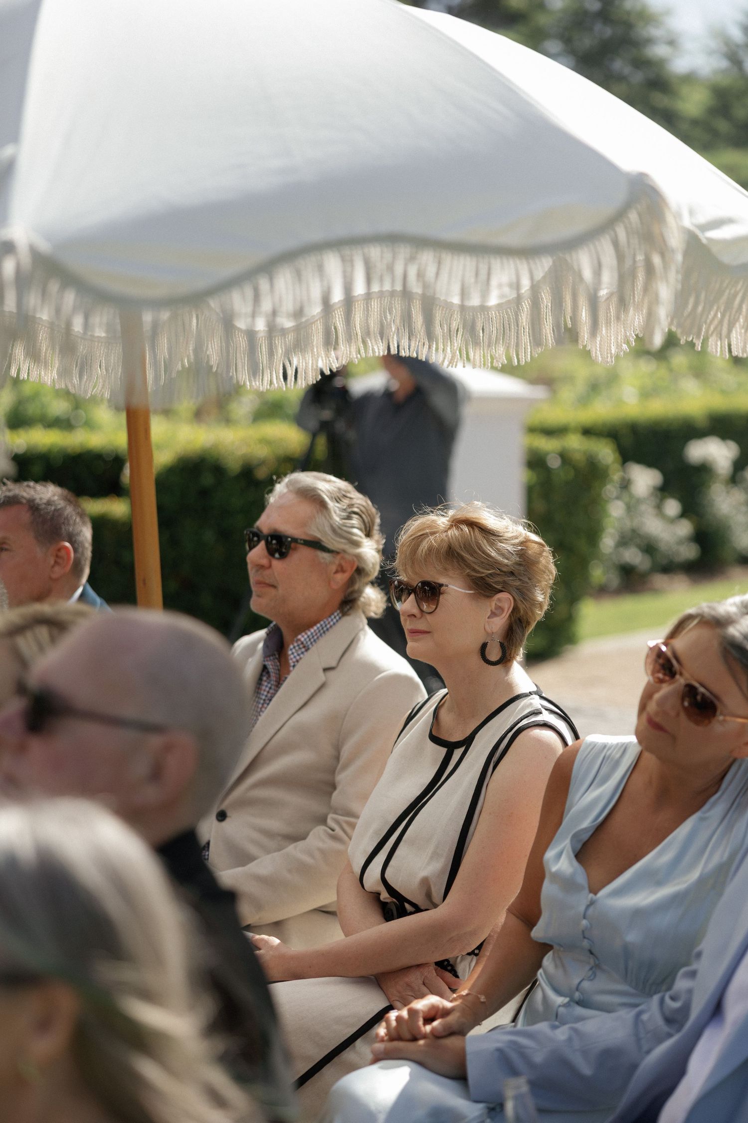 Elegantly dressed guests wearing sunglasses seated outdoors under a white fringed umbrella at a garden event.