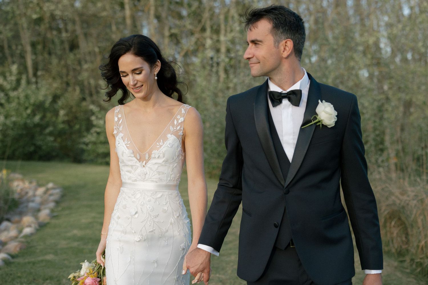 Bride in lace gown and groom in black tuxedo holding hands outdoors in garden.