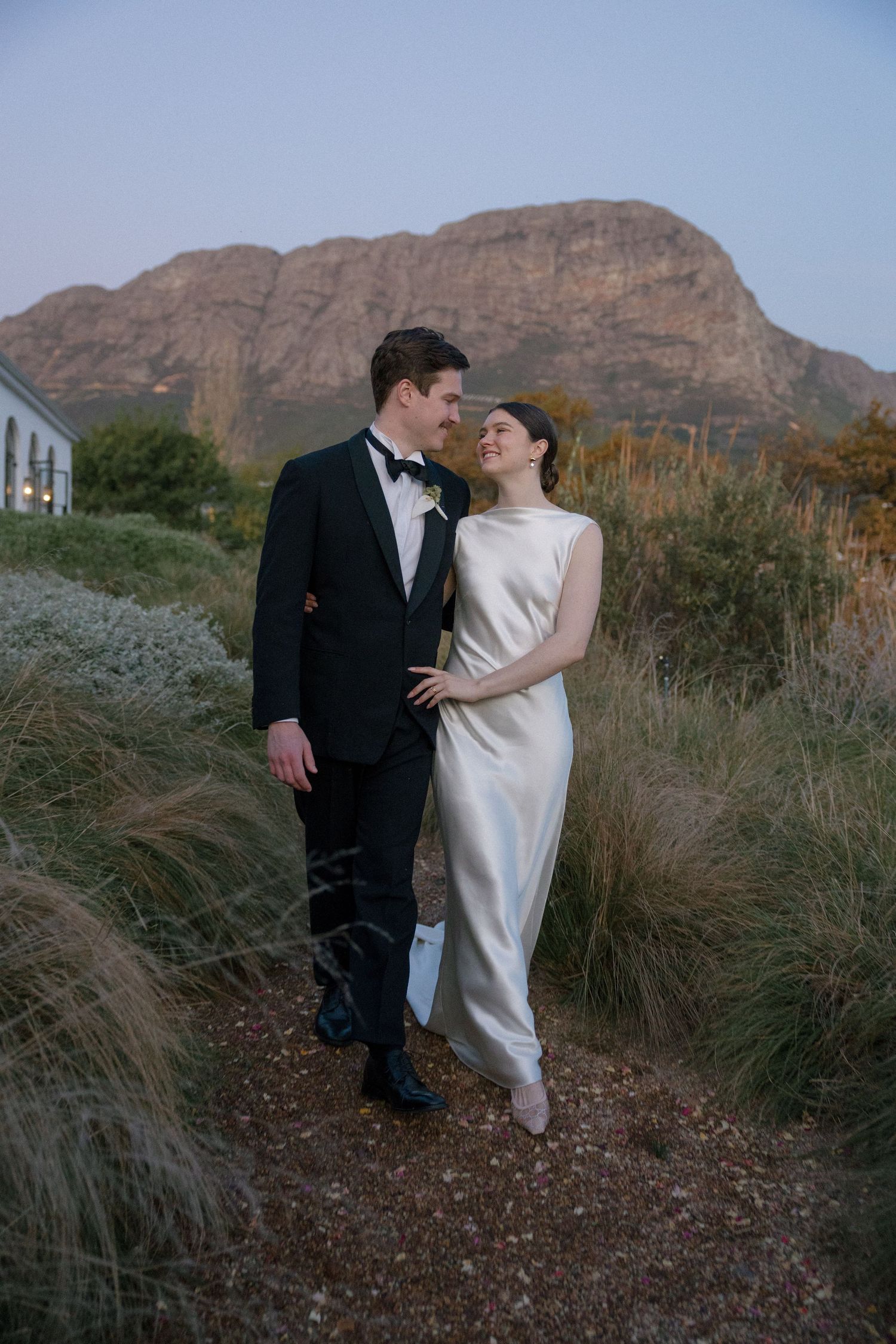 Couple gazes lovingly at each other on a gravel path surrounded by fynbos with mountain backdrop.