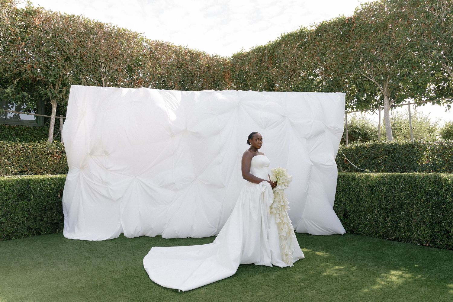 Elegant bride in strapless ball gown with long train poses before white draped backdrop in garden.