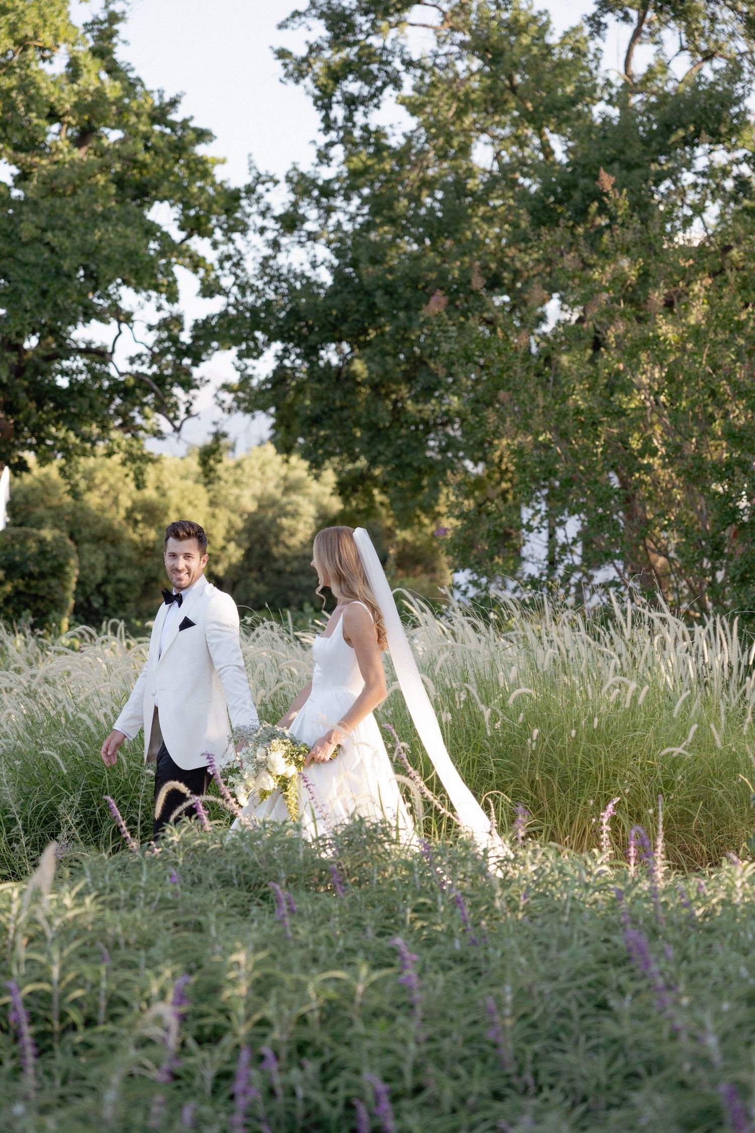 Bride and groom walking through tall grass meadow with wildflowers and trees at golden hour wedding.