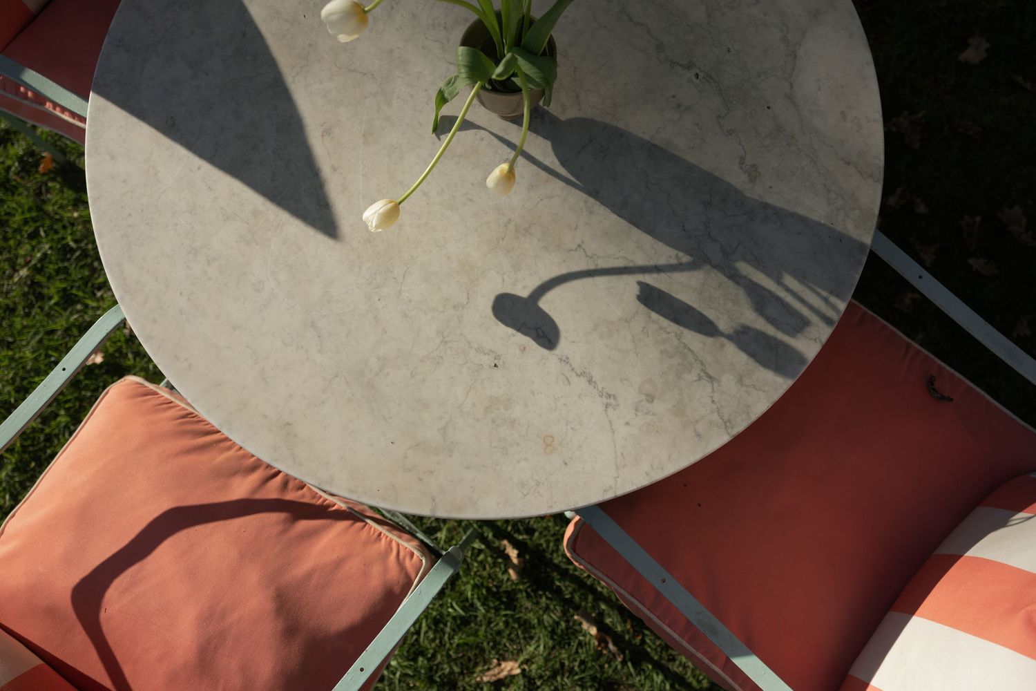 Aerial view of round stone patio table with white calla lilies casting shadows, flanked by chairs with coral cushions.