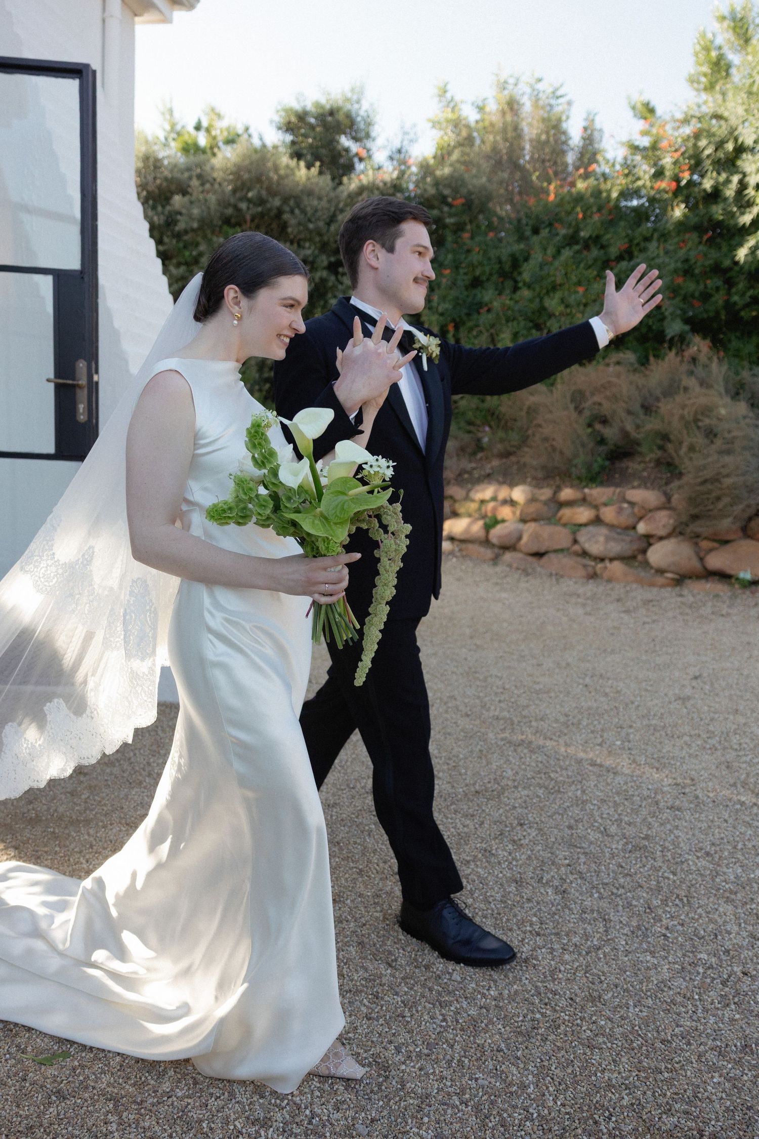 Bride and groom walking outdoors after wedding ceremony, groom waving joyfully while bride holds white floral bouquet.
