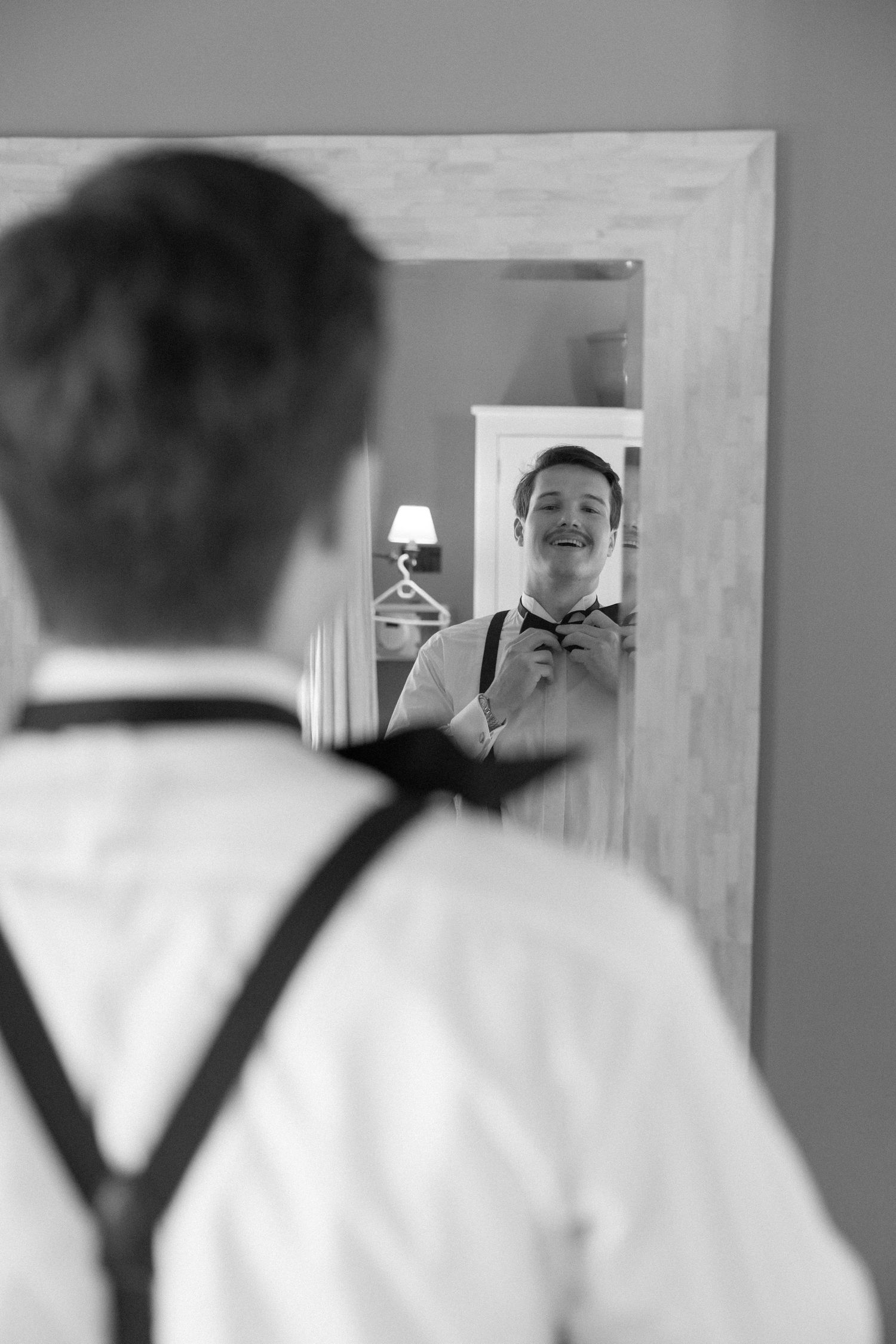 Groom smiling in mirror reflection while adjusting bow tie, wearing suspenders, black and white wedding photo.