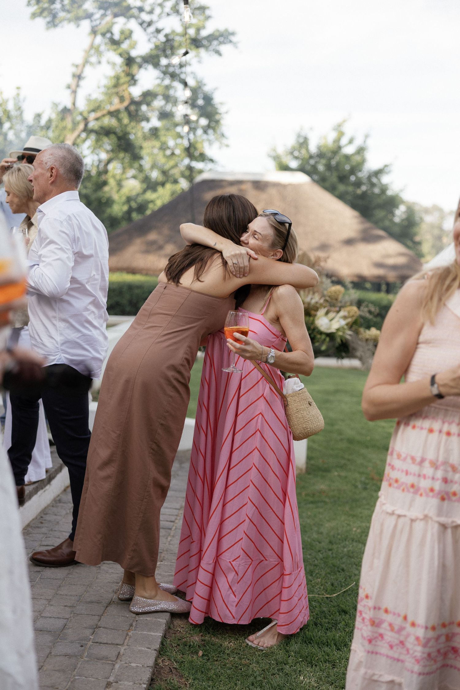 Two women in summer dresses share a warm hug at an outdoor garden party event.