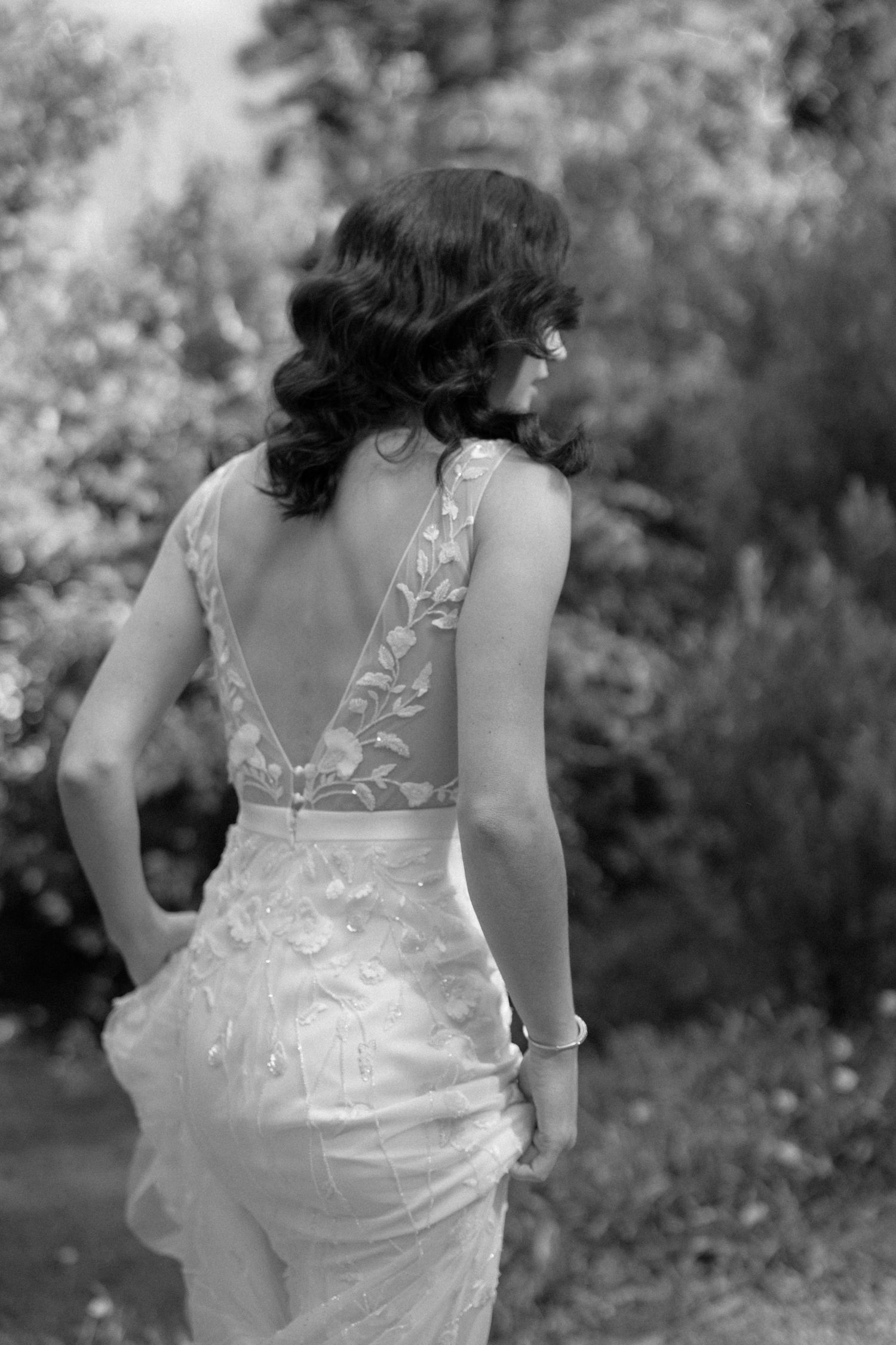 Black and white rear view of a bride in a low-back floral appliqué gown standing in a sunlit garden.