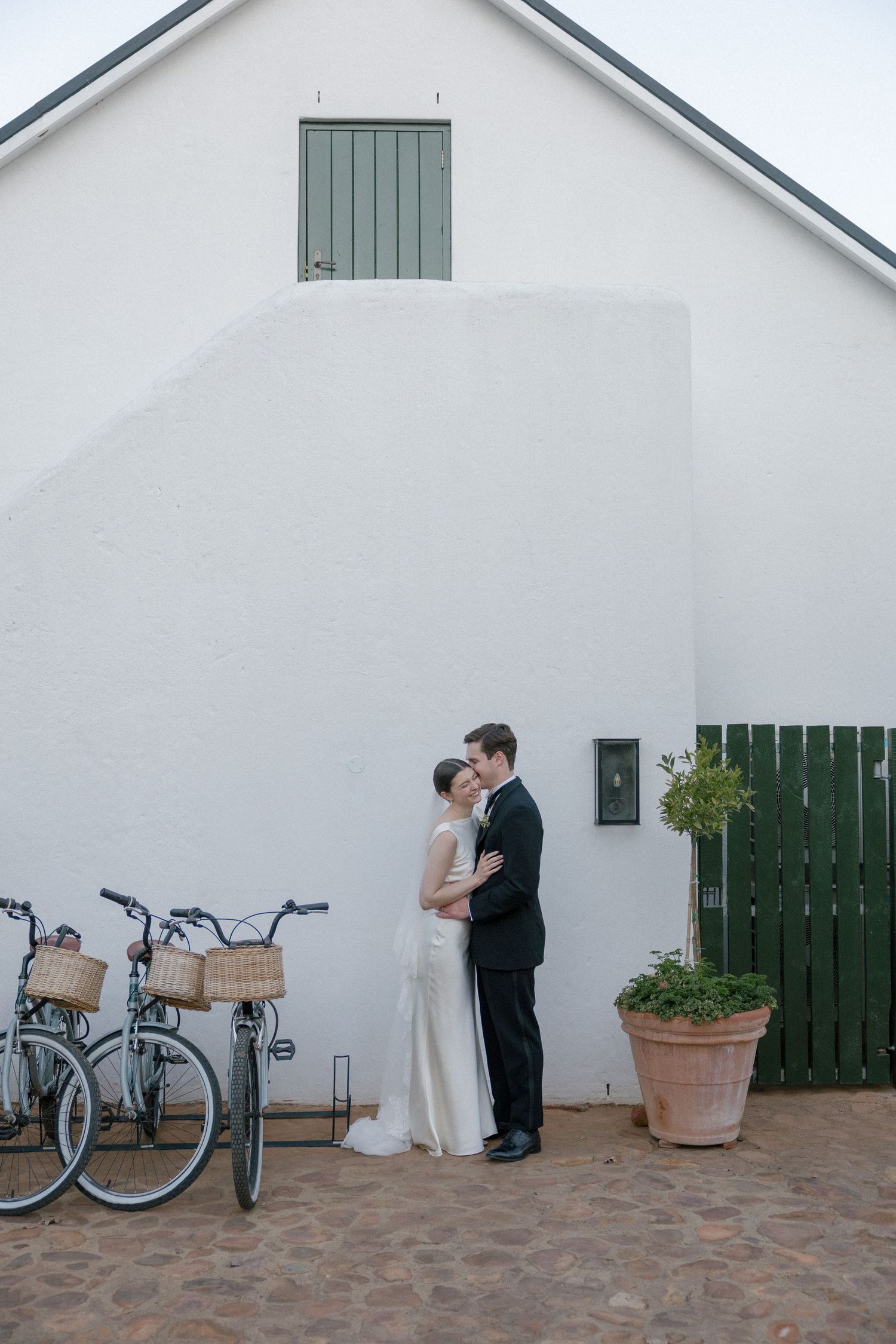 Bride and groom embracing beside a white barn wall with bicycles and a green gate.