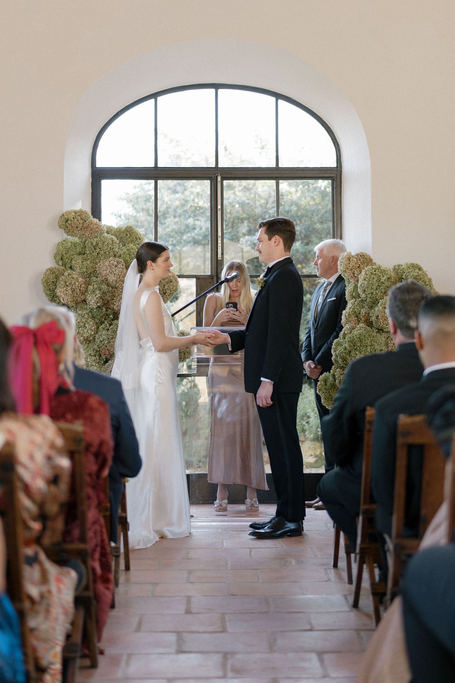 Bride and groom hold hands at altar flanked by tall hydrangea arrangements in sunlit chapel.