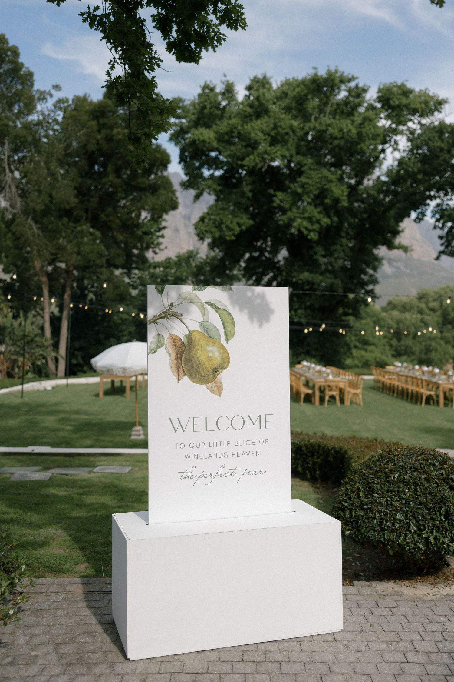 Elegant outdoor wedding welcome sign with pear illustration on white pedestal, garden venue with string lights and mountains.