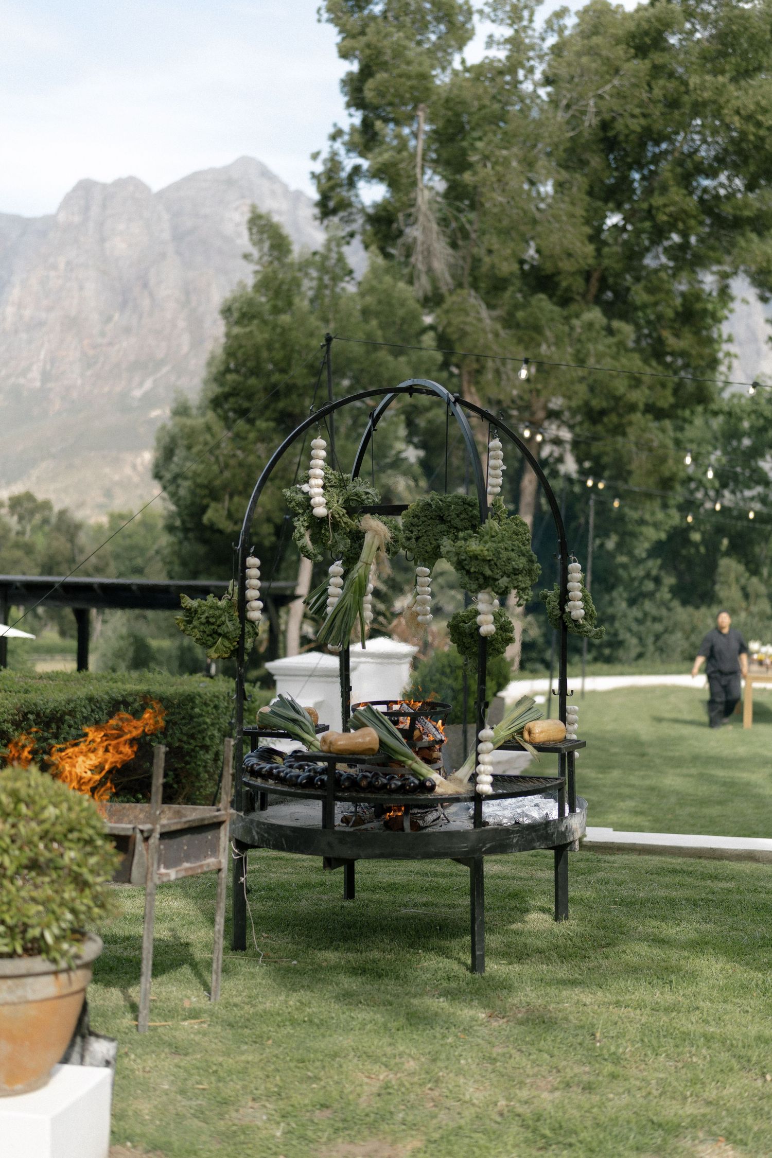 Elegant outdoor braai station with decorative arch, greenery, and string lights set against mountain backdrop.