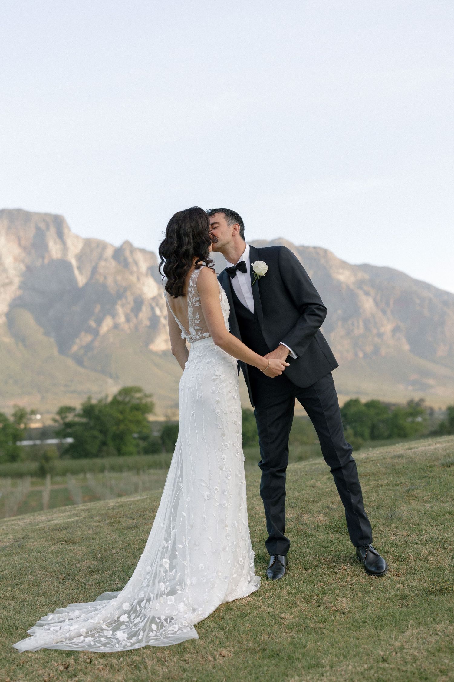 Groom in black tuxedo kissing bride in lace gown with sweeping mountain views behind them.
