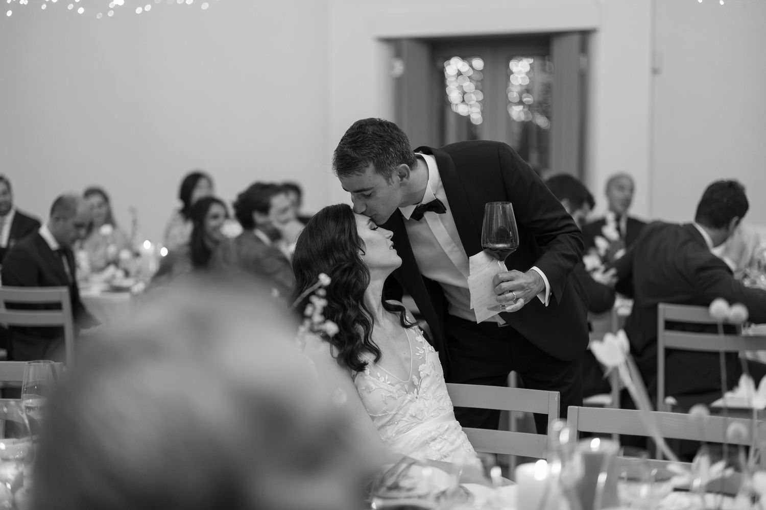 Bride and groom share a romantic kiss at wedding reception table in black and white photo.