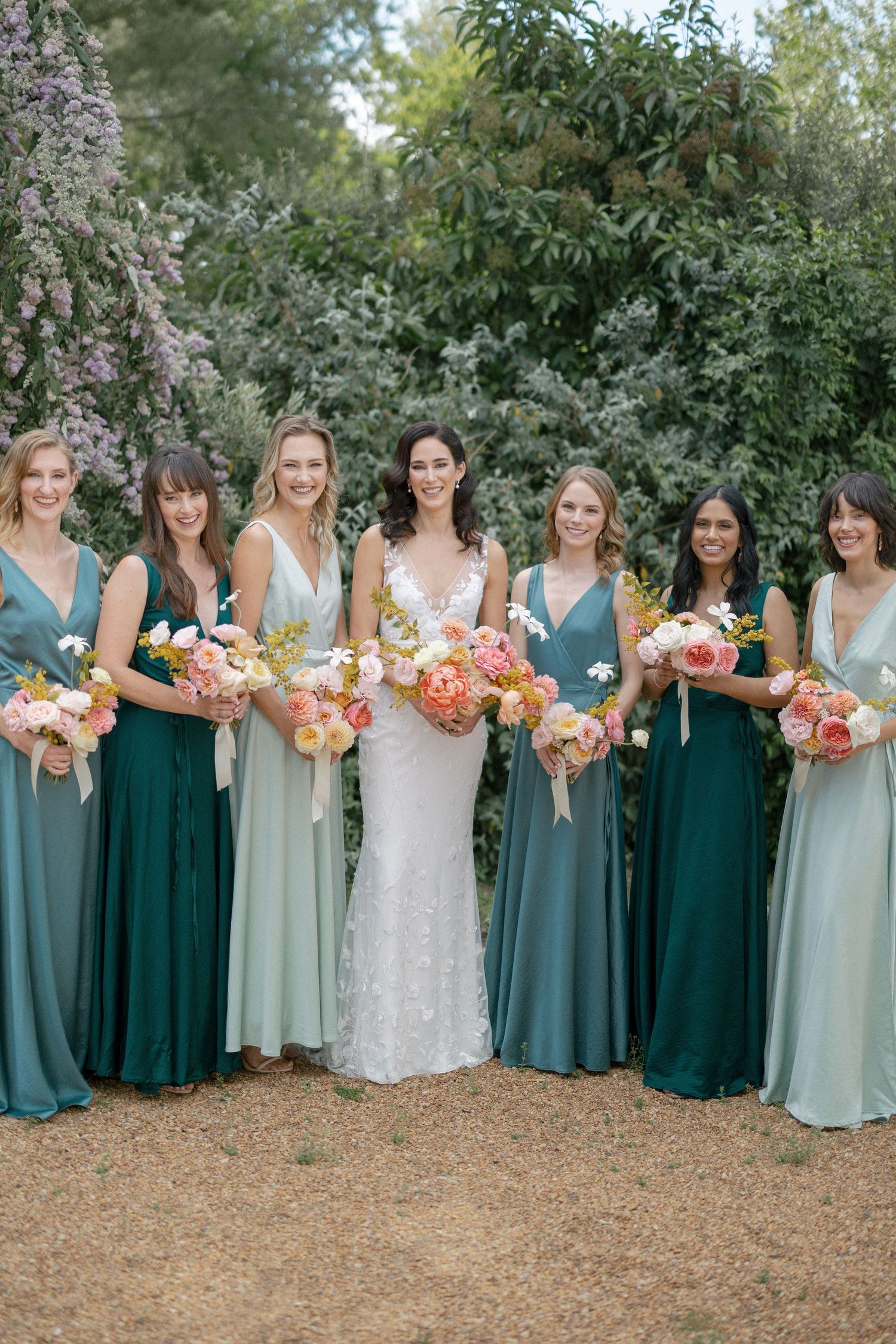 Bride in a fitted beaded gown poses with six bridesmaids in mismatched green dresses holding colorful floral bouquets.
