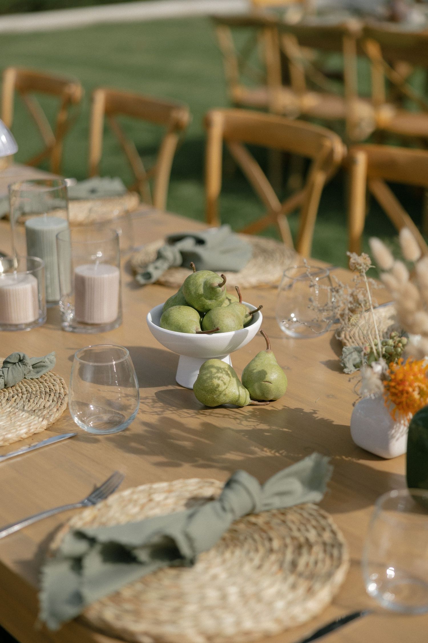 Outdoor wedding reception table with green pears, sage napkins, wicker placemats, candles, and wildflower arrangements.
