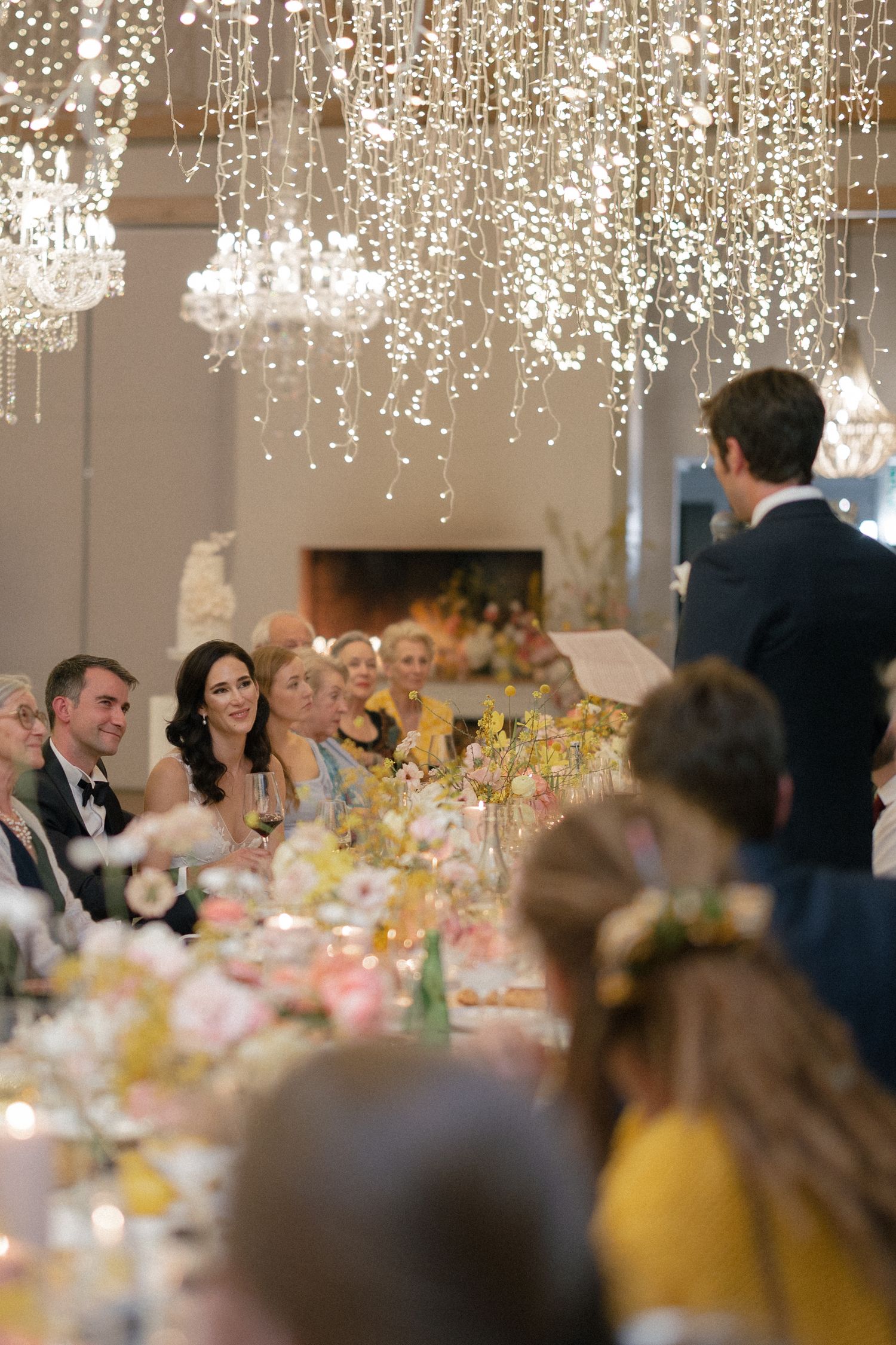 Wedding guests smile at head table beneath cascading fairy lights at elegant reception.
