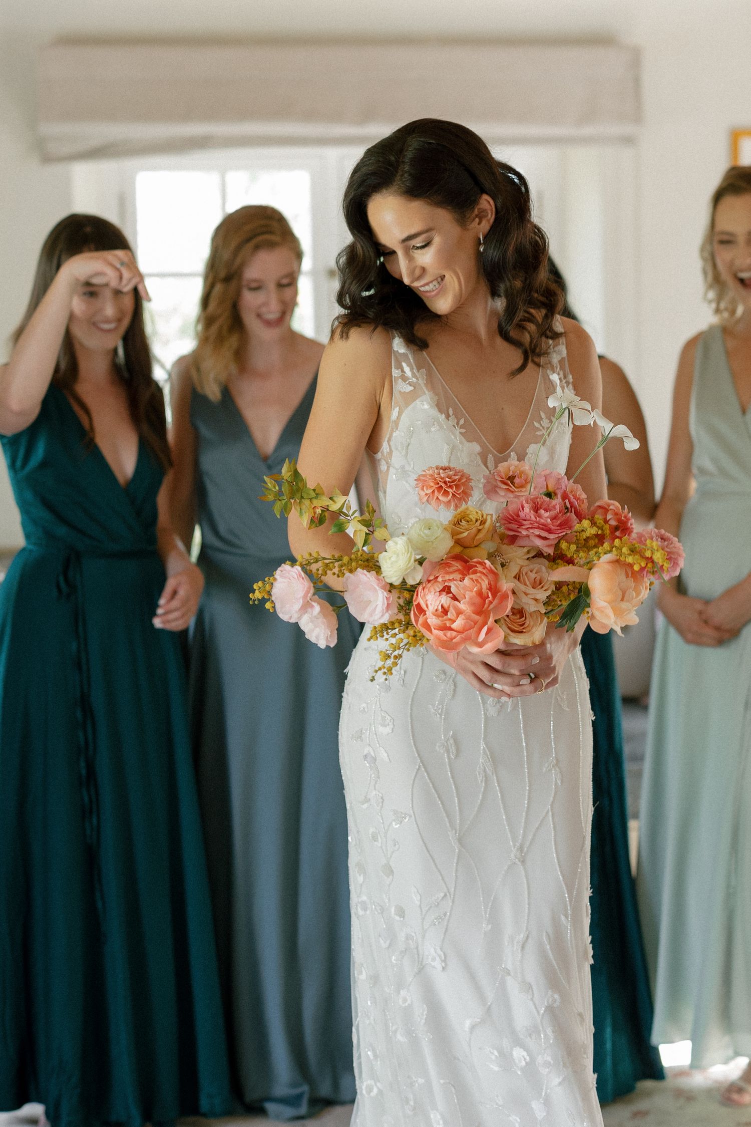Smiling bride in a floral beaded gown holds a vibrant coral and peach bouquet surrounded by bridesmaids in teal dresses.